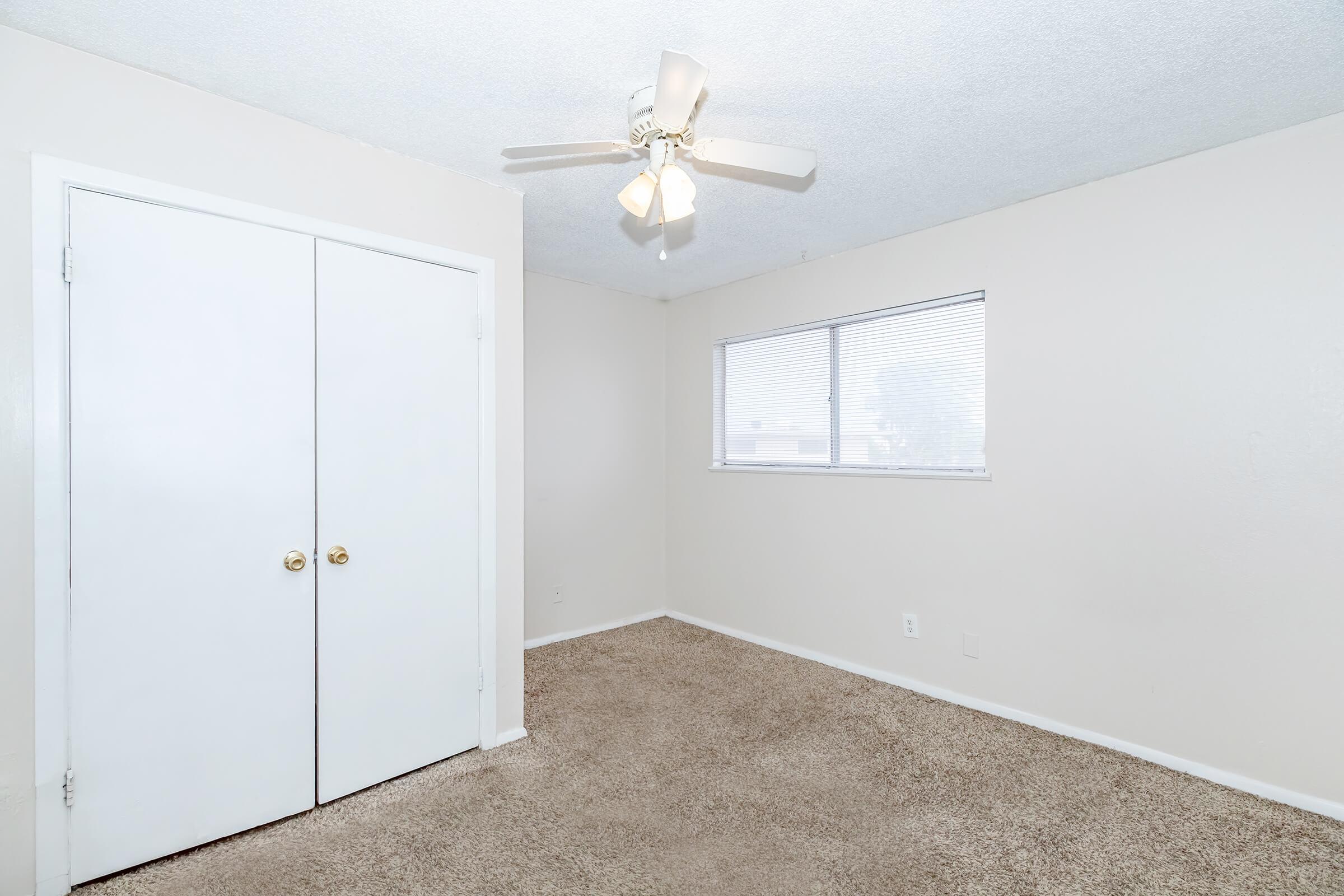 A well-lit empty bedroom featuring beige walls and plush carpeting. The room includes a ceiling fan, a window with blinds, and a double closet with white doors. The overall ambiance is clean and inviting, suitable for a new occupant to personalize.