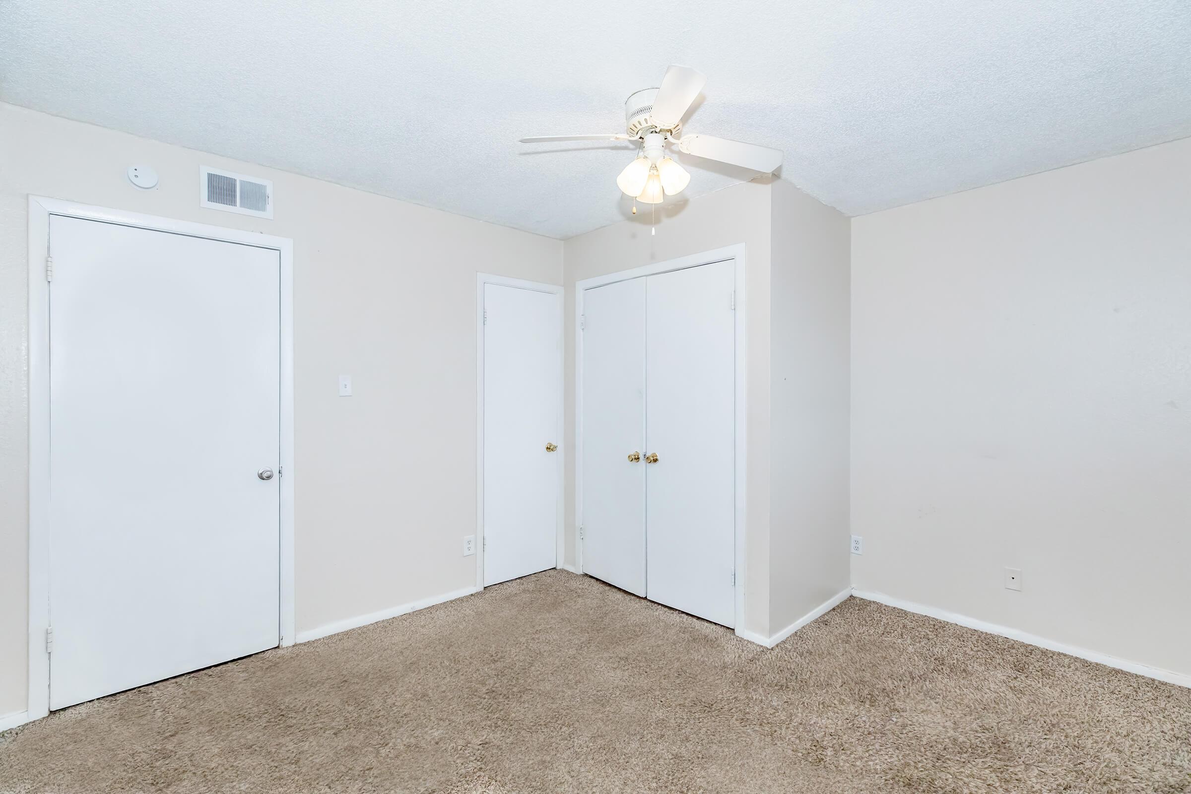 A simple, empty room featuring beige walls, a ceiling fan with lights, and plush carpet. Two closed closet doors are on the right, while a door leading outside is on the left. The space appears well-lit and minimalistic, suitable for various design possibilities.
