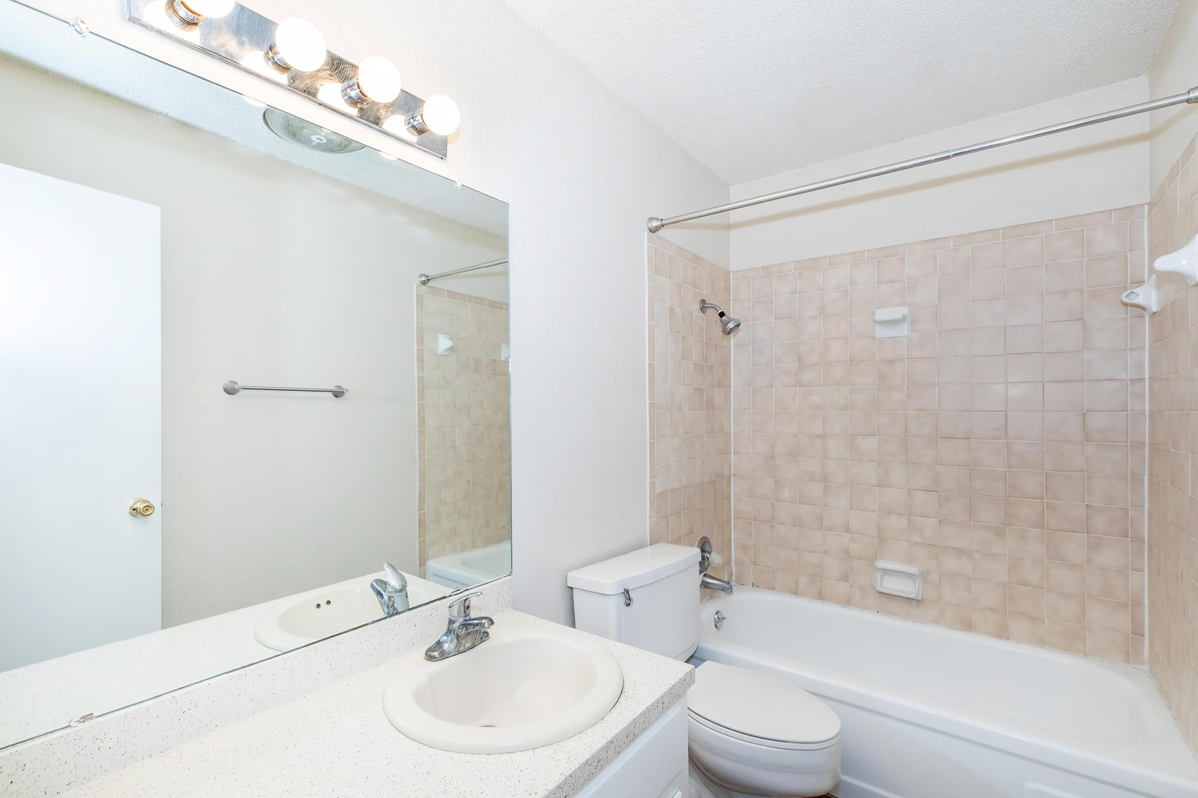 A well-lit bathroom featuring a bathtub with a shower, a white toilet, and a sink with a mirror above it. The walls are tiled in a beige color, and there is a towel rack mounted on one wall. The overall design is clean and simple, creating a spacious appearance.