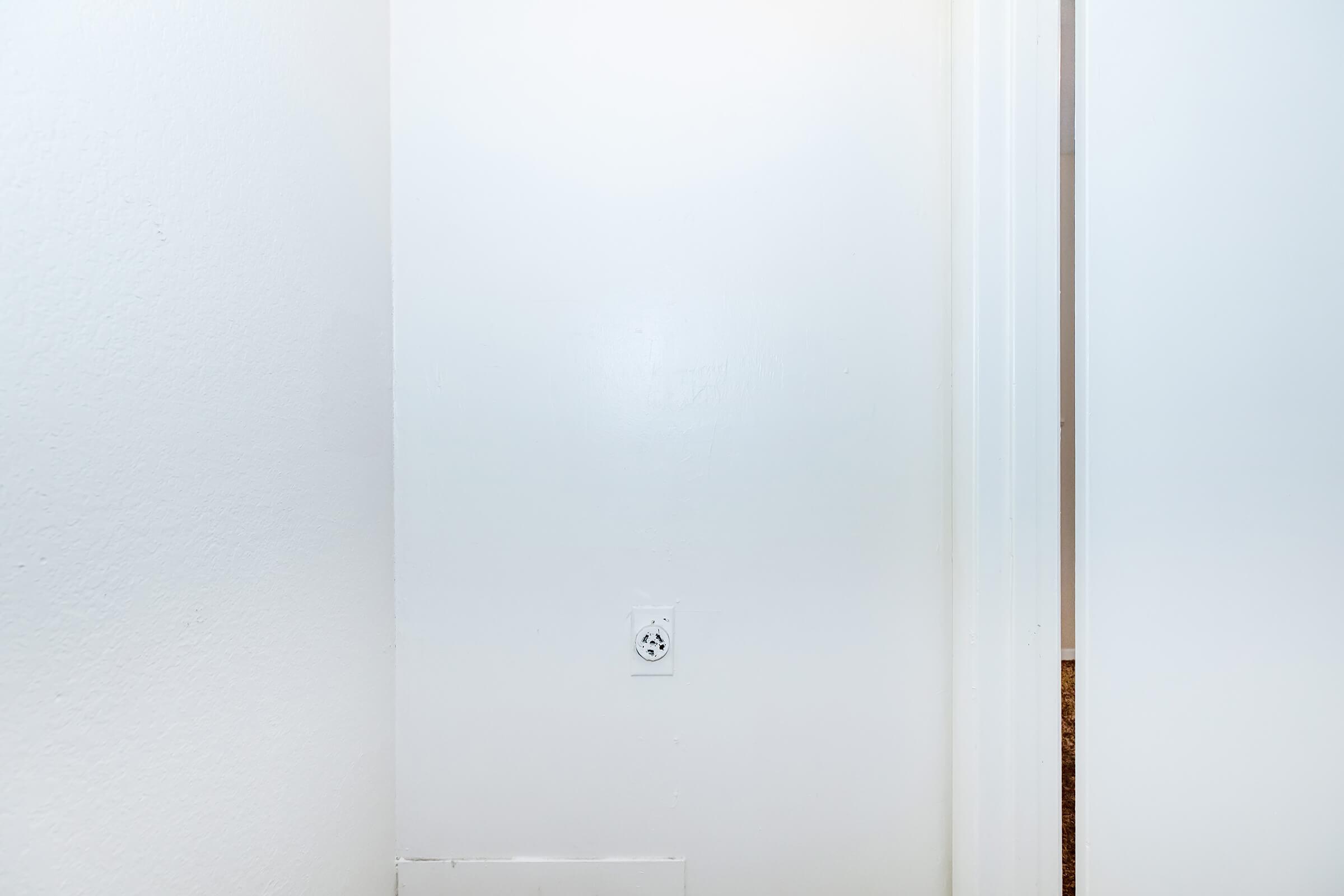 A plain white wall with a visible electrical outlet. On the right, there is a partially open white door leading to another room. The area is brightly lit, emphasizing the simple and clean aesthetic of the space.