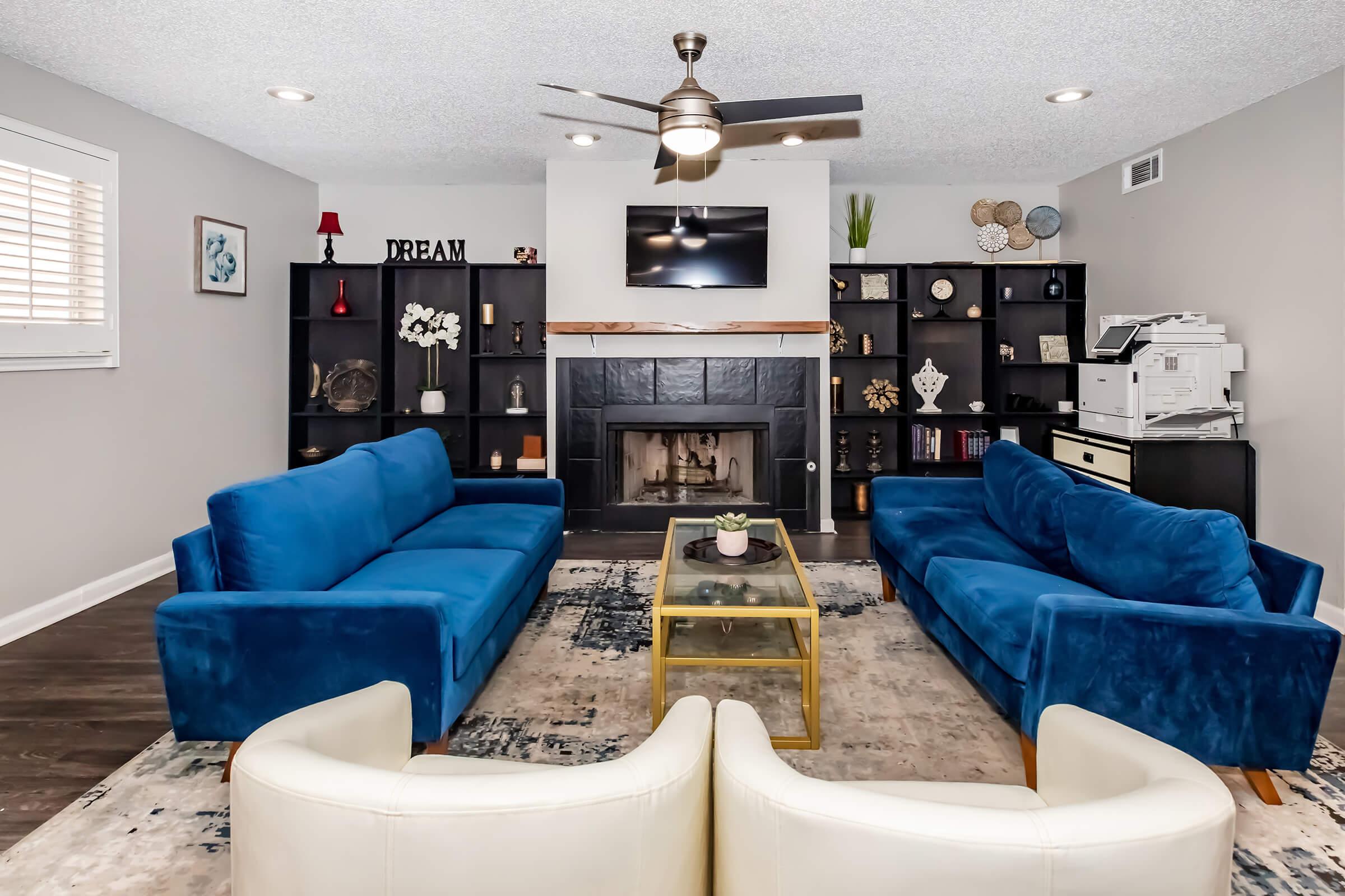 A cozy living room featuring two blue sofas and a white armchair. A coffee table sits on a decorative rug in front of the seating area. The wall has a mounted TV above a black fireplace, with built-in shelves displaying decor items and a printer on the side. Light gray walls and wood flooring complete the space.