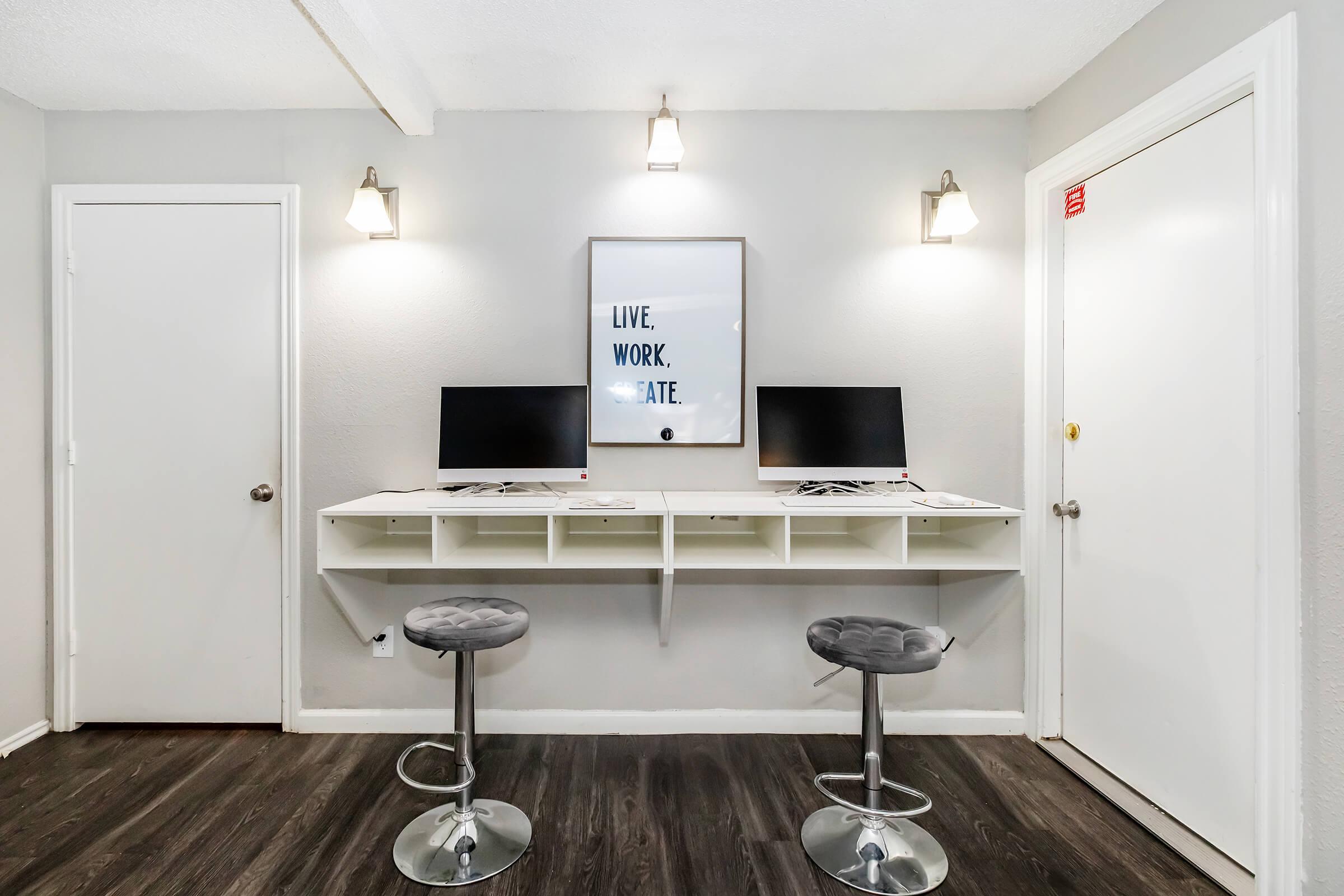 A modern workspace featuring two computer monitors on a white wall-mounted desk. The area is well-lit with two wall sconces, and a motivational poster reading "LIVE, WORK, CREATE" is displayed above the desk. There are two stylish gray bar stools in front of the desk, and a white door is visible to the right.