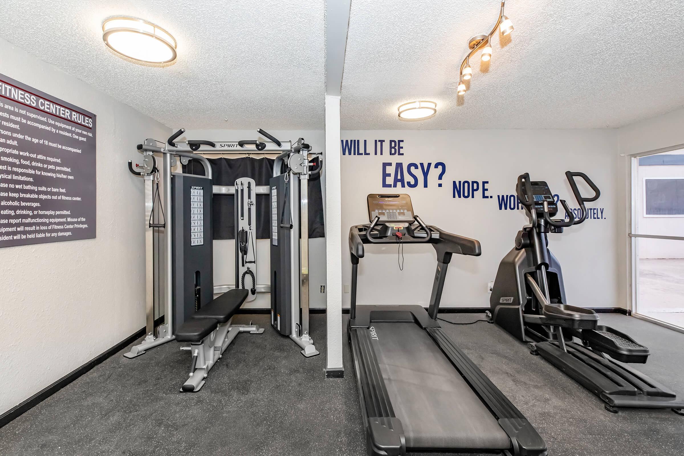 A fitness center with workout equipment including a multi-station gym machine, a treadmill, and an elliptical machine. The area has a motivational wall quote: "WILL IT BE EASY? NOPE. WORK HARD." Light fixtures illuminate the room, which features a clean and modern design.