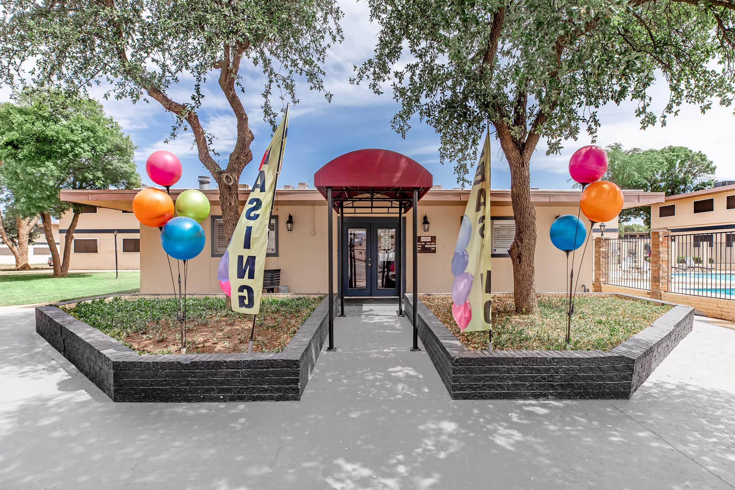 Entrance to a building with a red awning, flanked by large colorful balloons and banners that read "CASINO." Green grass and trees surround the area, with a visible outdoor pool in the background. The scene is bright and inviting, suggesting a welcoming atmosphere.