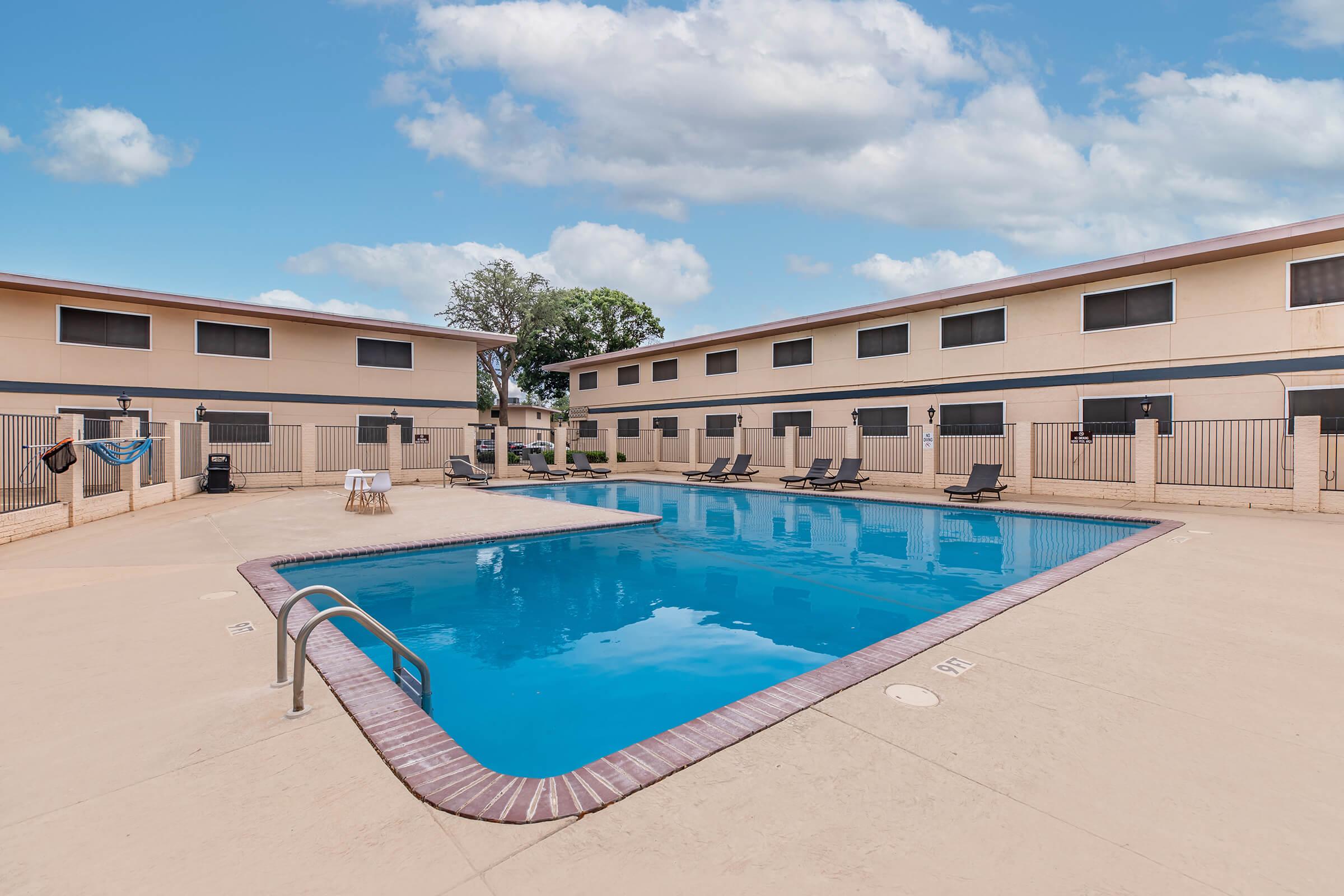 A clear blue swimming pool surrounded by a tan deck and lounge chairs, with two buildings in the background featuring multiple windows. The sky is bright with fluffy clouds, creating a relaxed outdoor atmosphere ideal for leisure.