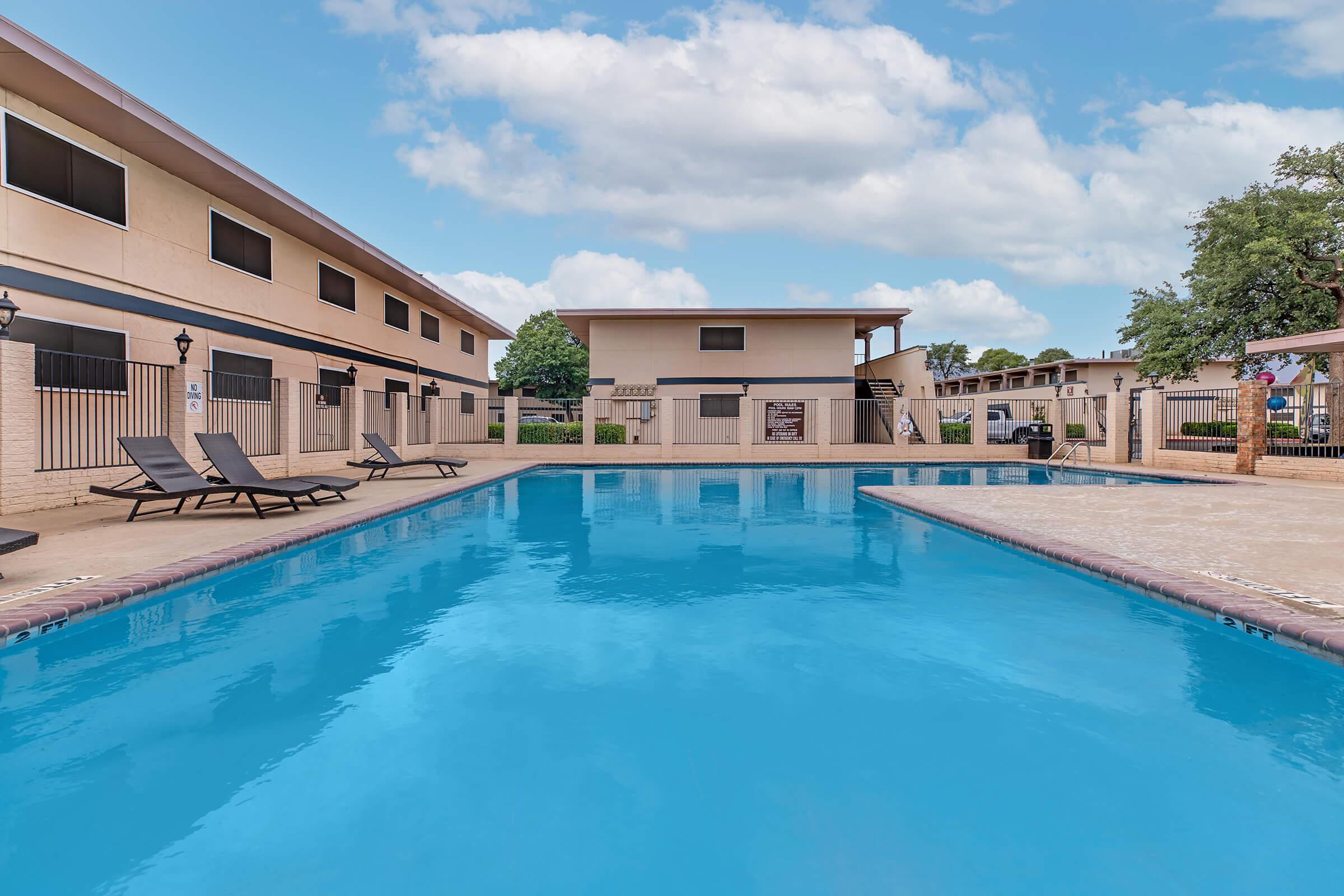 A spacious outdoor swimming pool surrounded by lounge chairs, with modern apartment buildings in the background. The sky is partly cloudy, and the area is well-maintained, inviting relaxation and enjoyment.