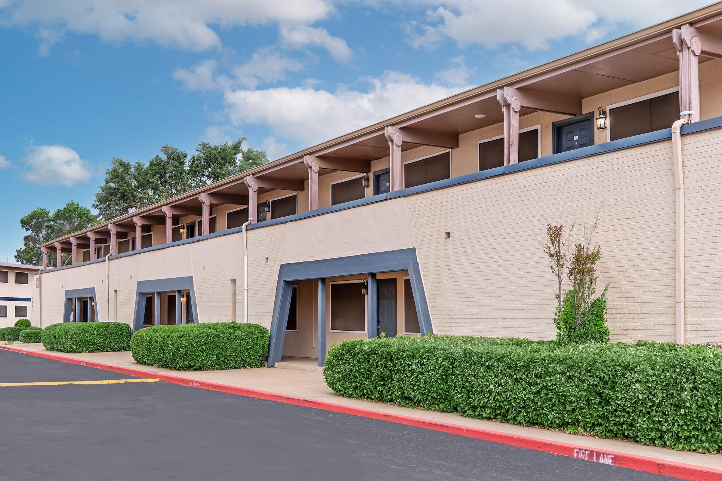 A low-rise building with multiple units features a light-colored exterior and blue accents. The structure has large windows, and neatly trimmed bushes line the walkway. The sky above is partly cloudy, adding a bright and inviting atmosphere to the scene.