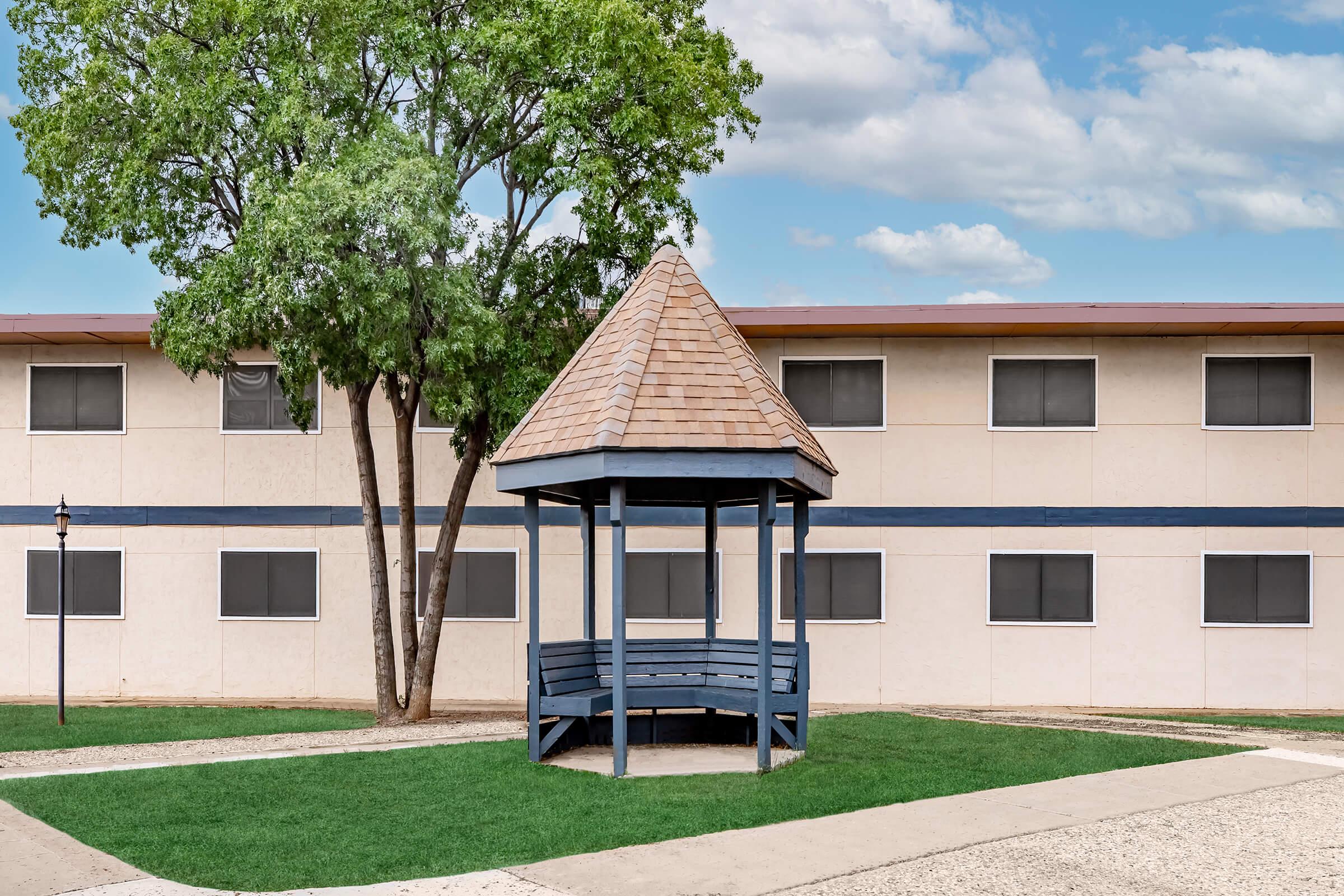 A blue gazebo with a shingled roof stands on a grassy area in front of a beige building featuring multiple windows. A tree is positioned near the gazebo, and the sky above is partly cloudy. The scene conveys a peaceful outdoor space.