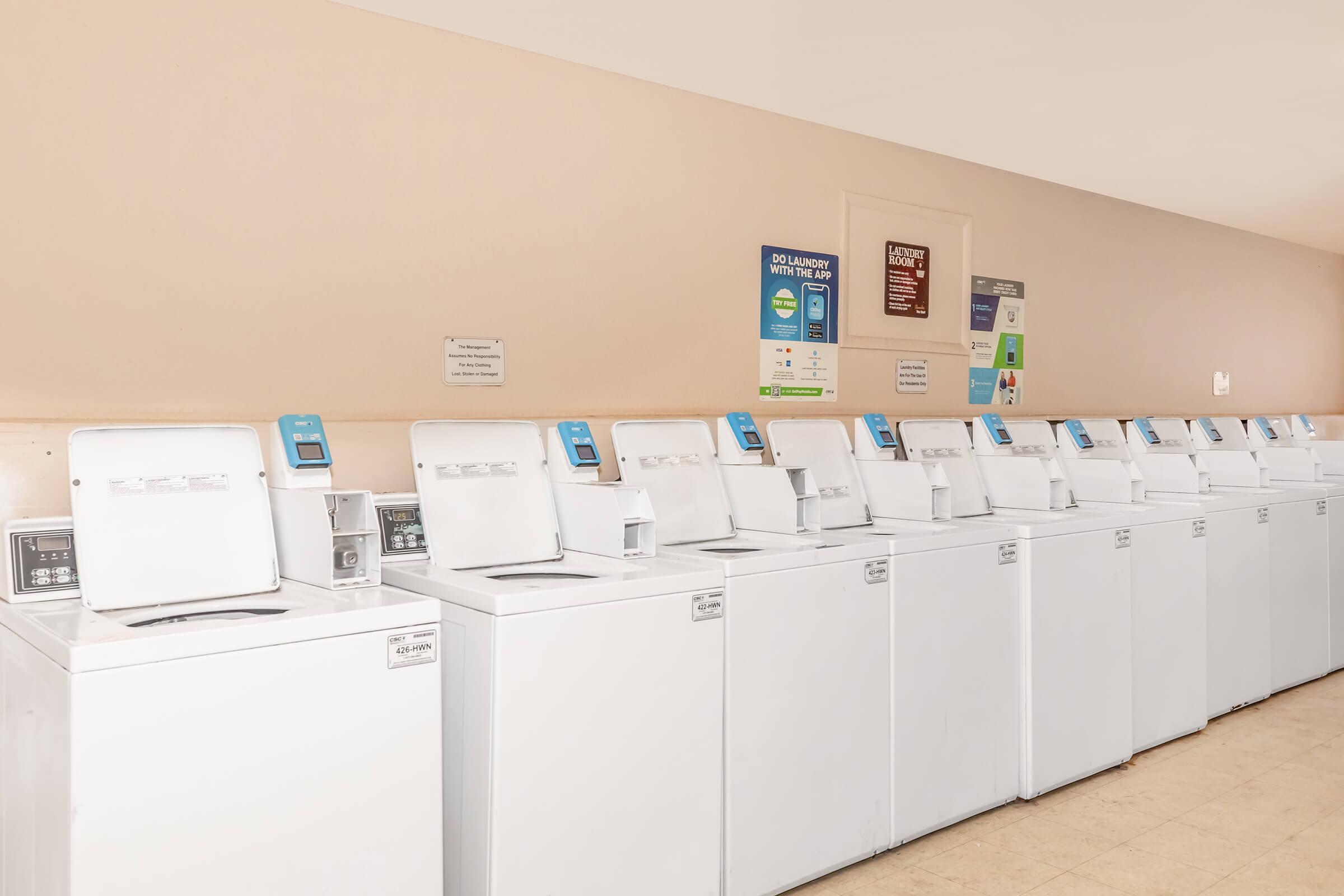 A row of modern washing machines in a laundry room. The machines are white and have digital controls, with signs on the wall providing instructions and laundry policies. The floor is tiled, and the overall ambiance is clean and tidy, suitable for communal laundry use.