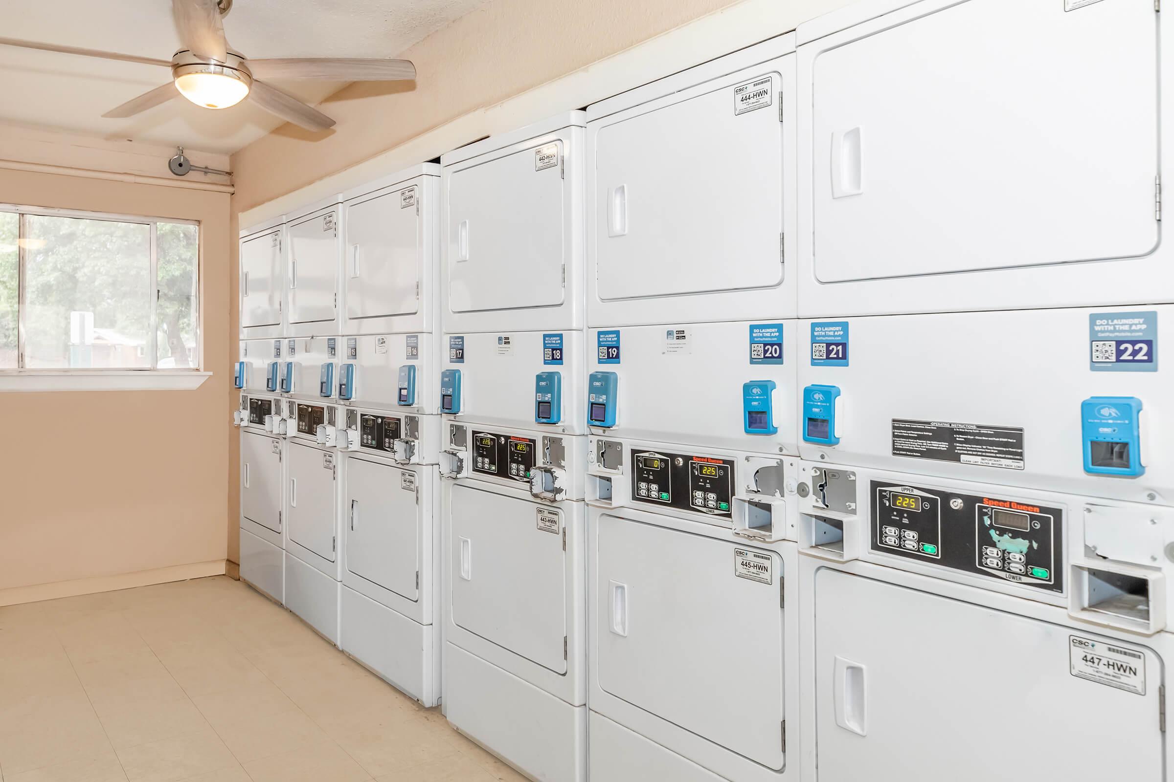 A clean laundry room featuring several white washing and drying machines in rows. Each machine has a digital display and a blue payment system. The room is well-lit with a ceiling fan, and a window allows natural light to enter, creating a bright and inviting atmosphere.