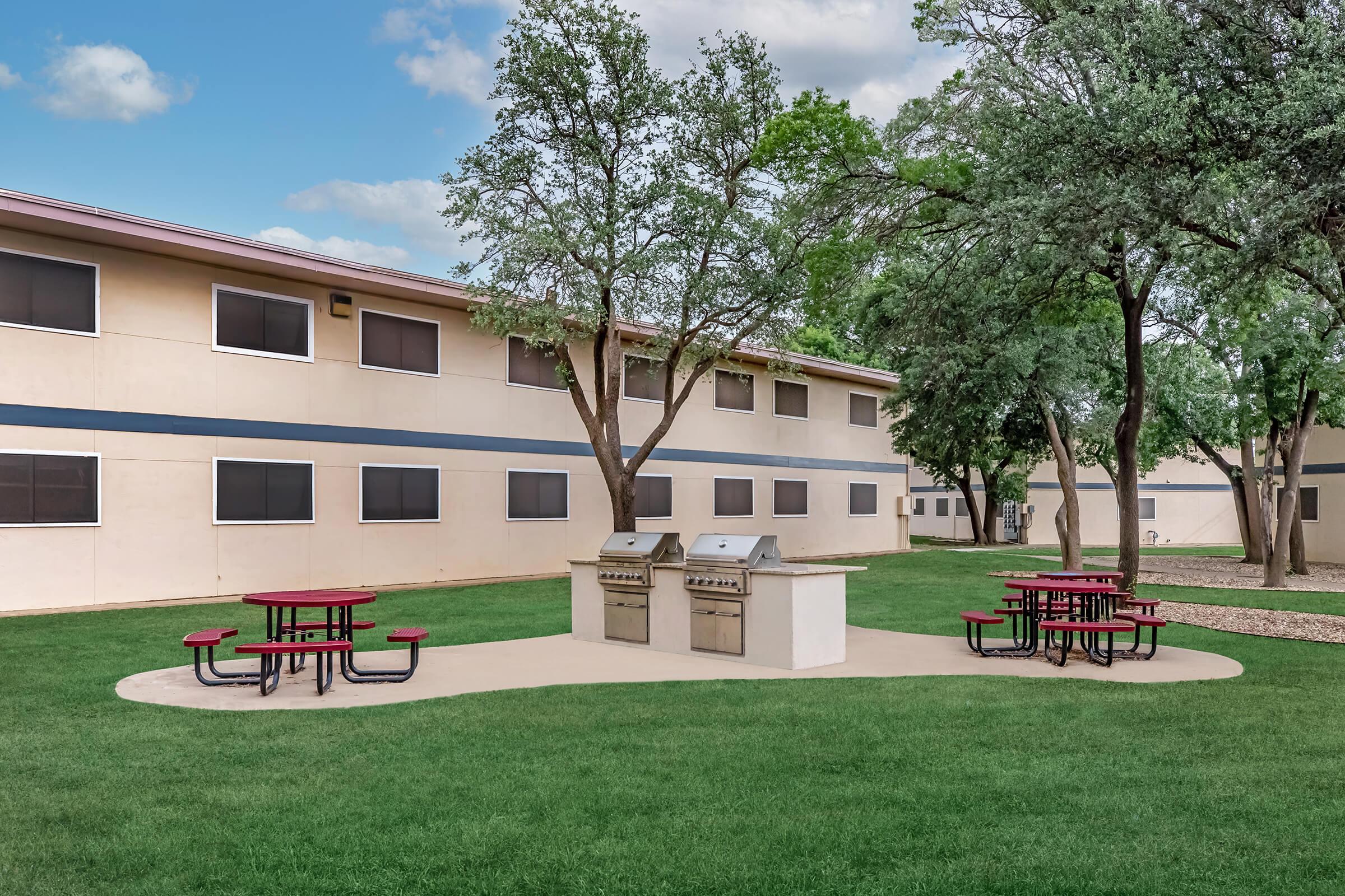 A grassy area featuring two barbecue grills and red picnic tables, surrounded by trees. In the background, there is a multi-story building with multiple windows. The scene has a clear blue sky with fluffy clouds, creating a pleasant outdoor setting.