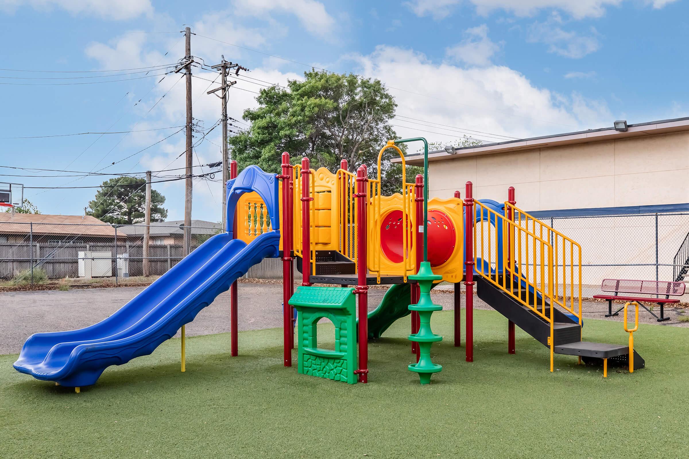 A colorful playground structure featuring blue and yellow slides, red and yellow climbing features, and a green playhouse. The area is surrounded by a fence and power lines, with a grassy surface underneath. The sky is clear with a few clouds.