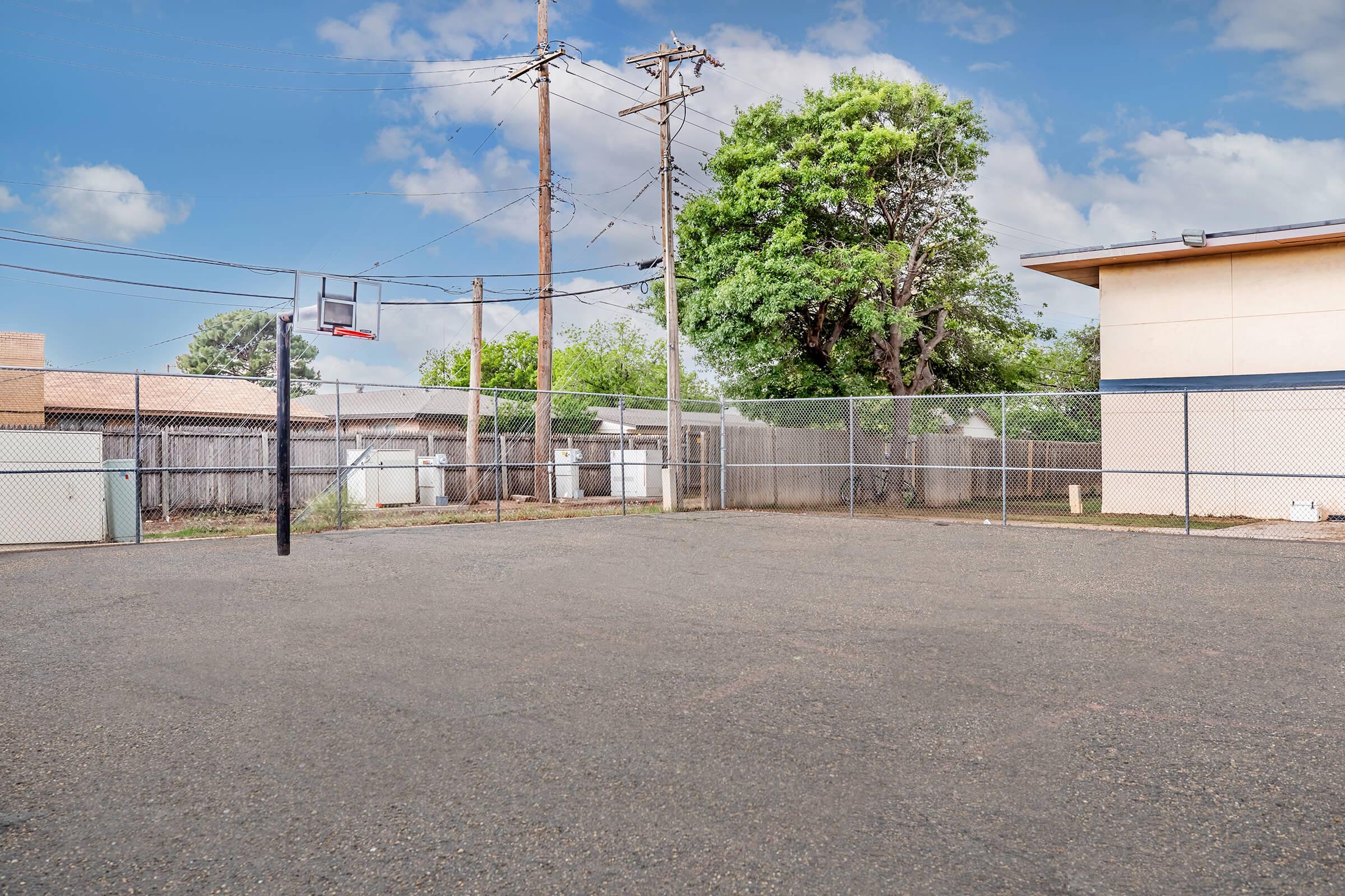 A view of an empty outdoor basketball court surrounded by a chain-link fence. In the background, there are power lines and trees, with a building visible on the right. The sky is blue with a few clouds, and the ground appears to be gravel or asphalt.