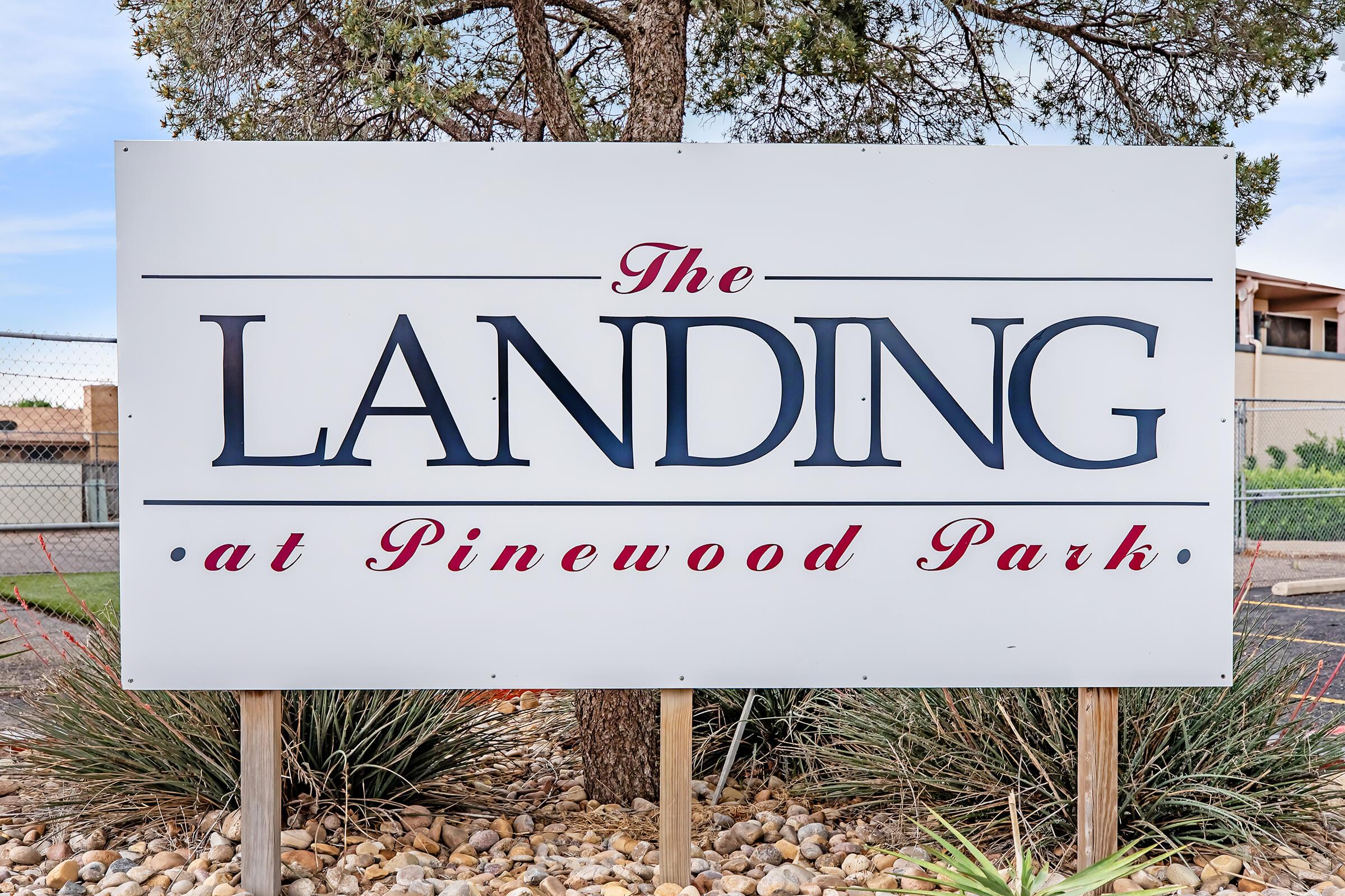 Sign for "The Landing at Pinewood Park," featuring the name prominently displayed in blue and red text, set among a landscaped area with rocks and shrubs. The background includes a clear sky and a fence.