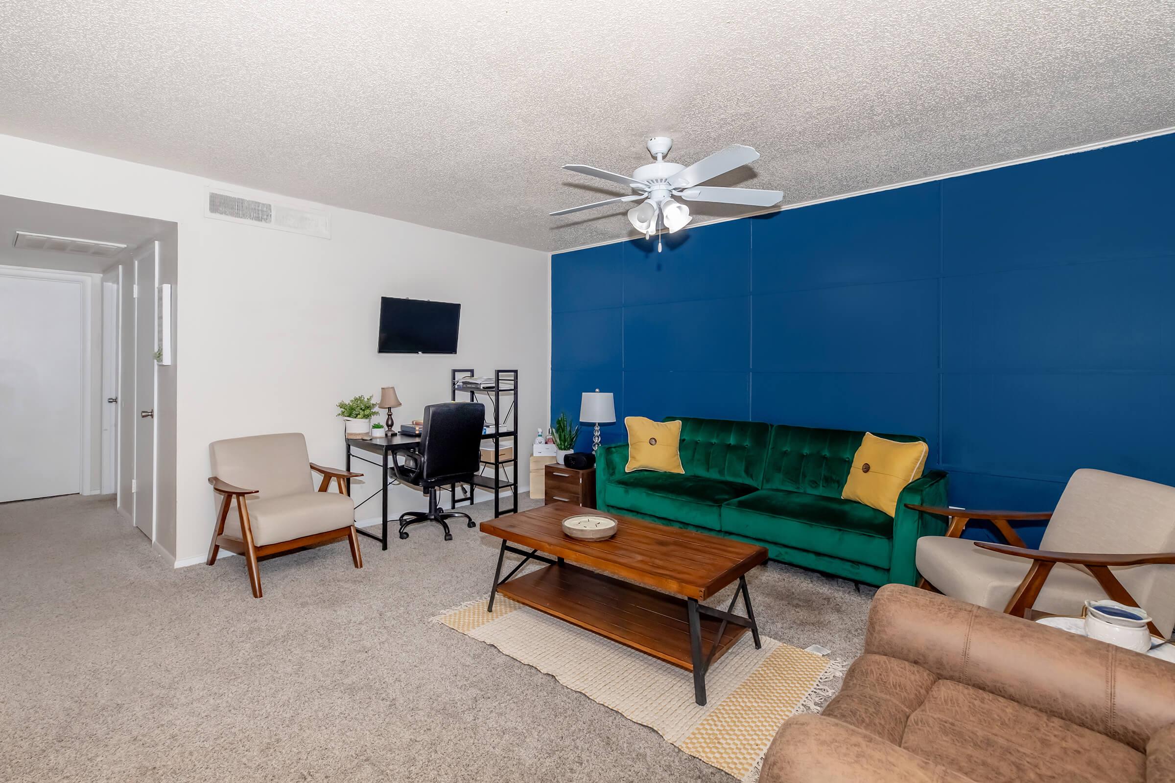 A modern living room featuring a green velvet sofa with yellow accents, a wooden coffee table, and beige armchairs. There's a ceiling fan, a wall-mounted TV, and a desk with an office chair against a blue accent wall. The flooring is carpeted, enhancing the cozy atmosphere.