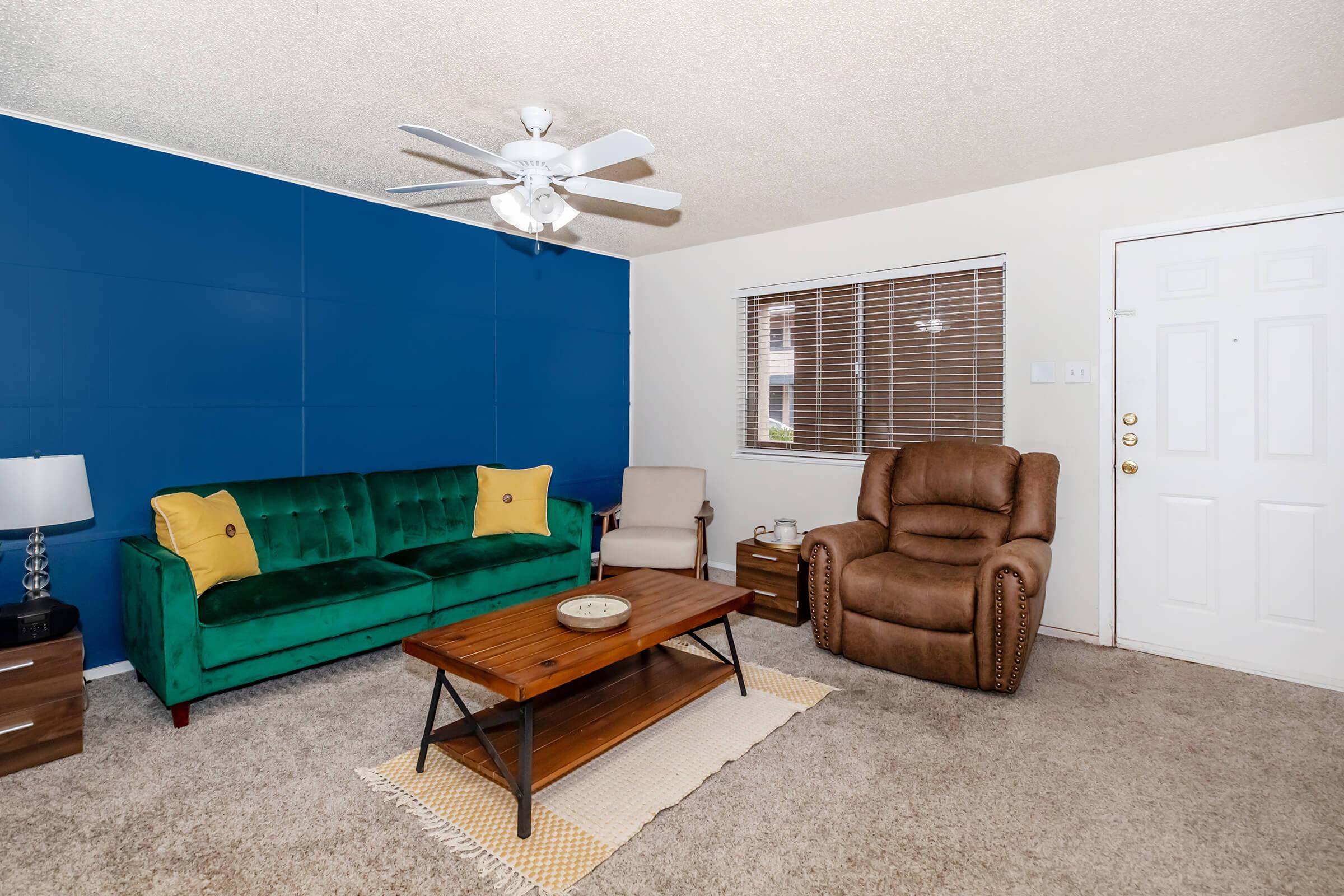 A cozy living room featuring a green velvet sofa with yellow accent pillows, a beige armchair, and a comfortable brown recliner. There's a wooden coffee table with a decorative item, a ceiling fan, and a blue accent wall. A window with blinds allows natural light, and the entrance door is visible on the right.