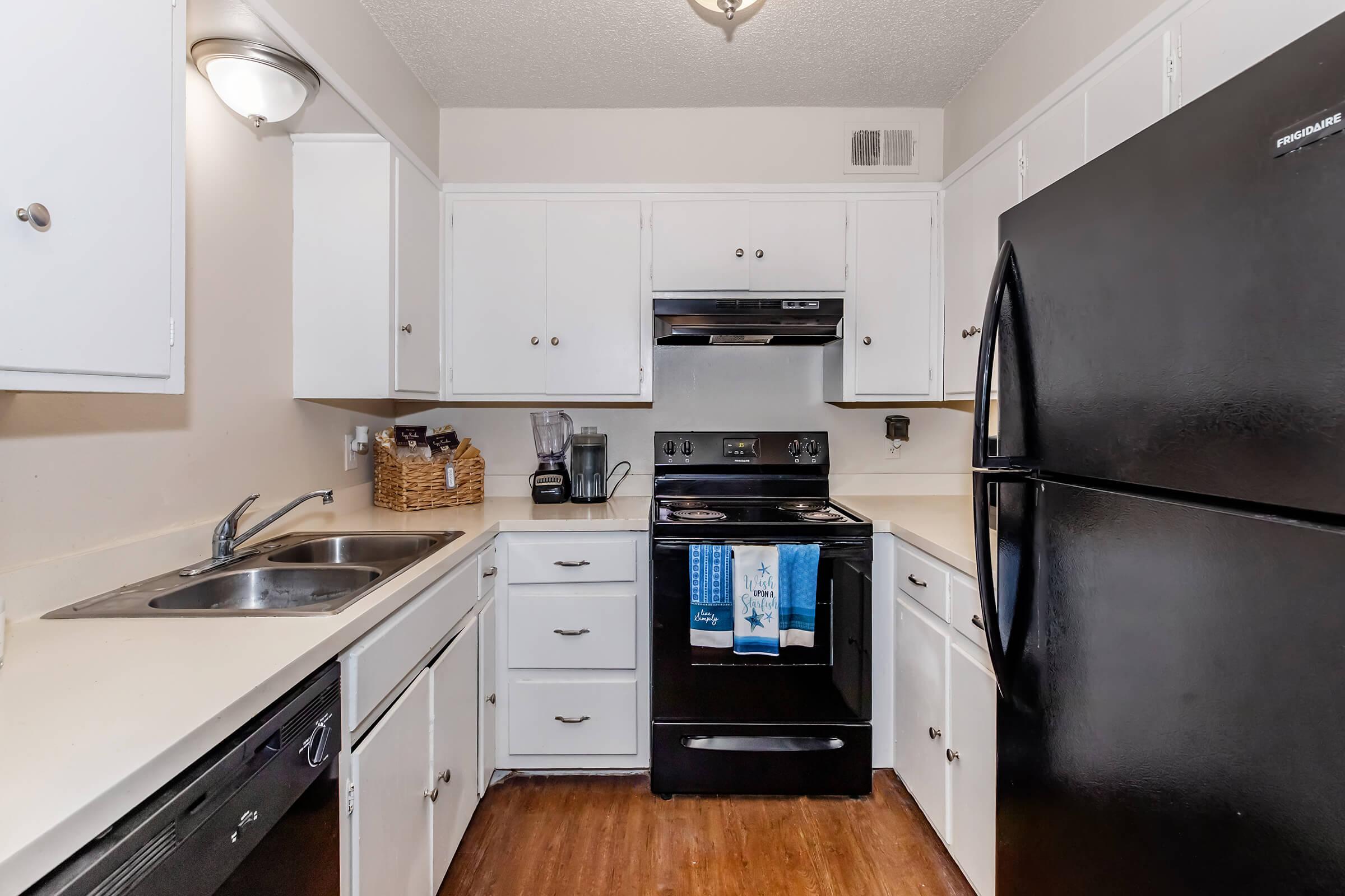 Compact kitchen featuring white cabinetry, black appliances including a refrigerator and oven, a sink with a double basin, and a countertop. A blender and dishware are also visible, along with a decorative basket on the counter. The flooring is a wood-like laminate, and the overall design is modern and functional.