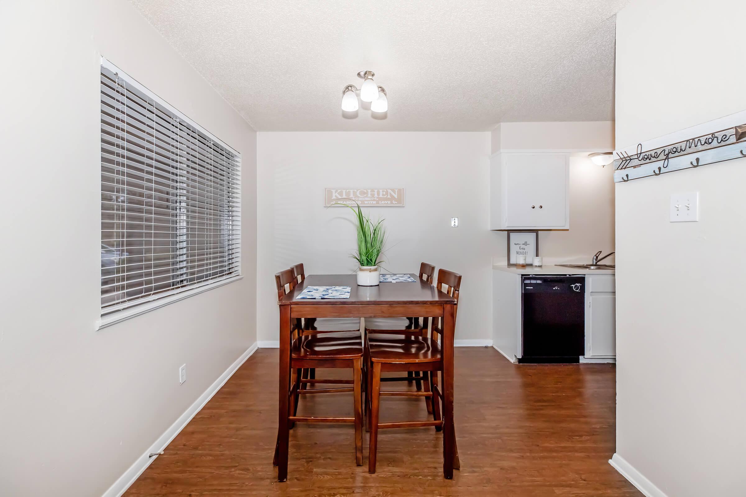 A bright, cozy kitchen with a wooden dining table and four chairs. The walls are painted light colors, and there are large blinds over the windows. A sign reading "KITCHEN" is displayed on the wall. The kitchen area is visible in the background, featuring white cabinetry and a black dishwasher.