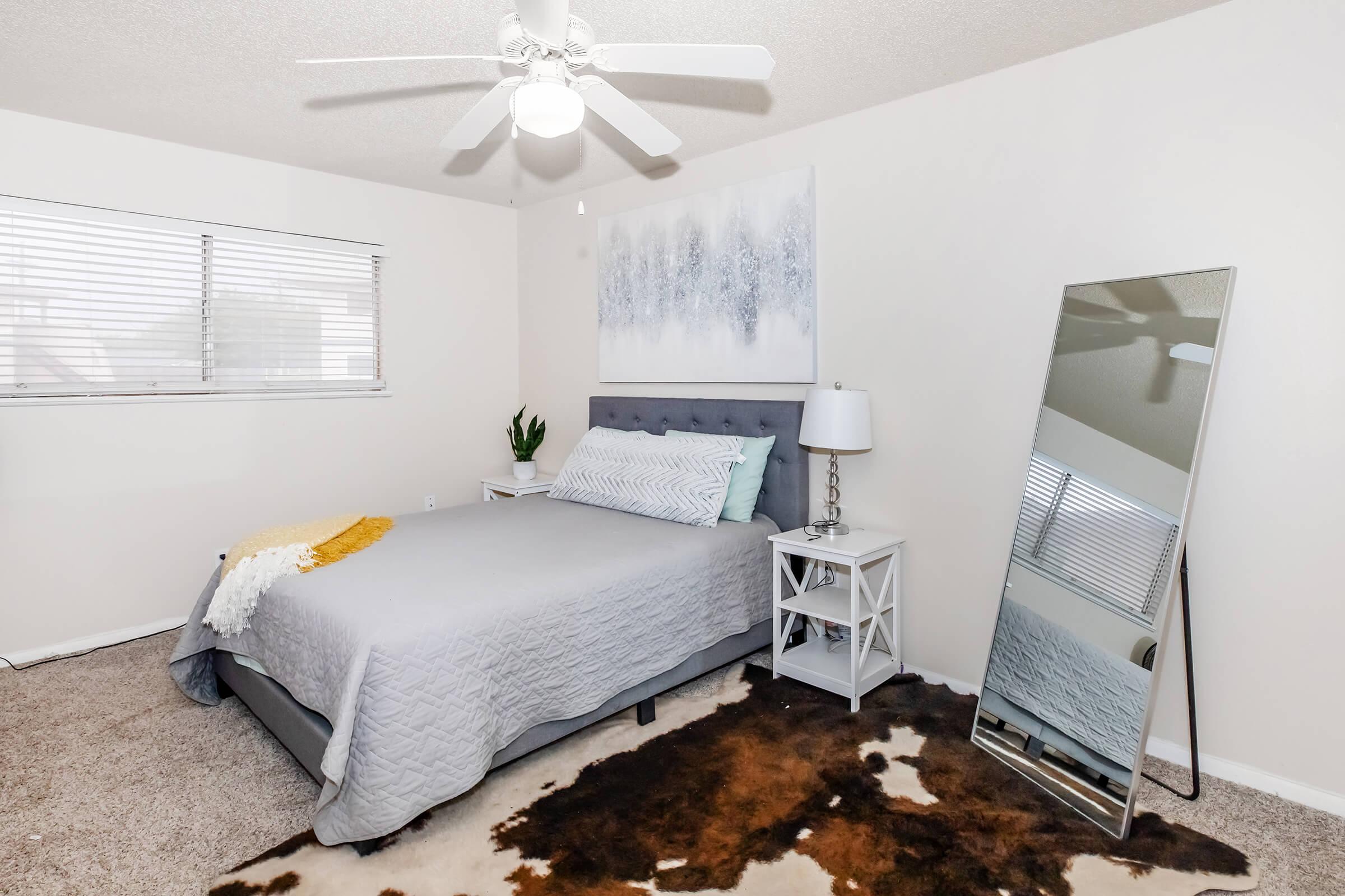 A cozy bedroom featuring a light gray bed with a textured blanket, a stylish lamp on a white bedside table, a large mirror, and a decorative piece of wall art. Natural light streams in through a window with blinds, complementing the neutral color scheme and inviting ambiance. A plush carpet adds warmth to the space.