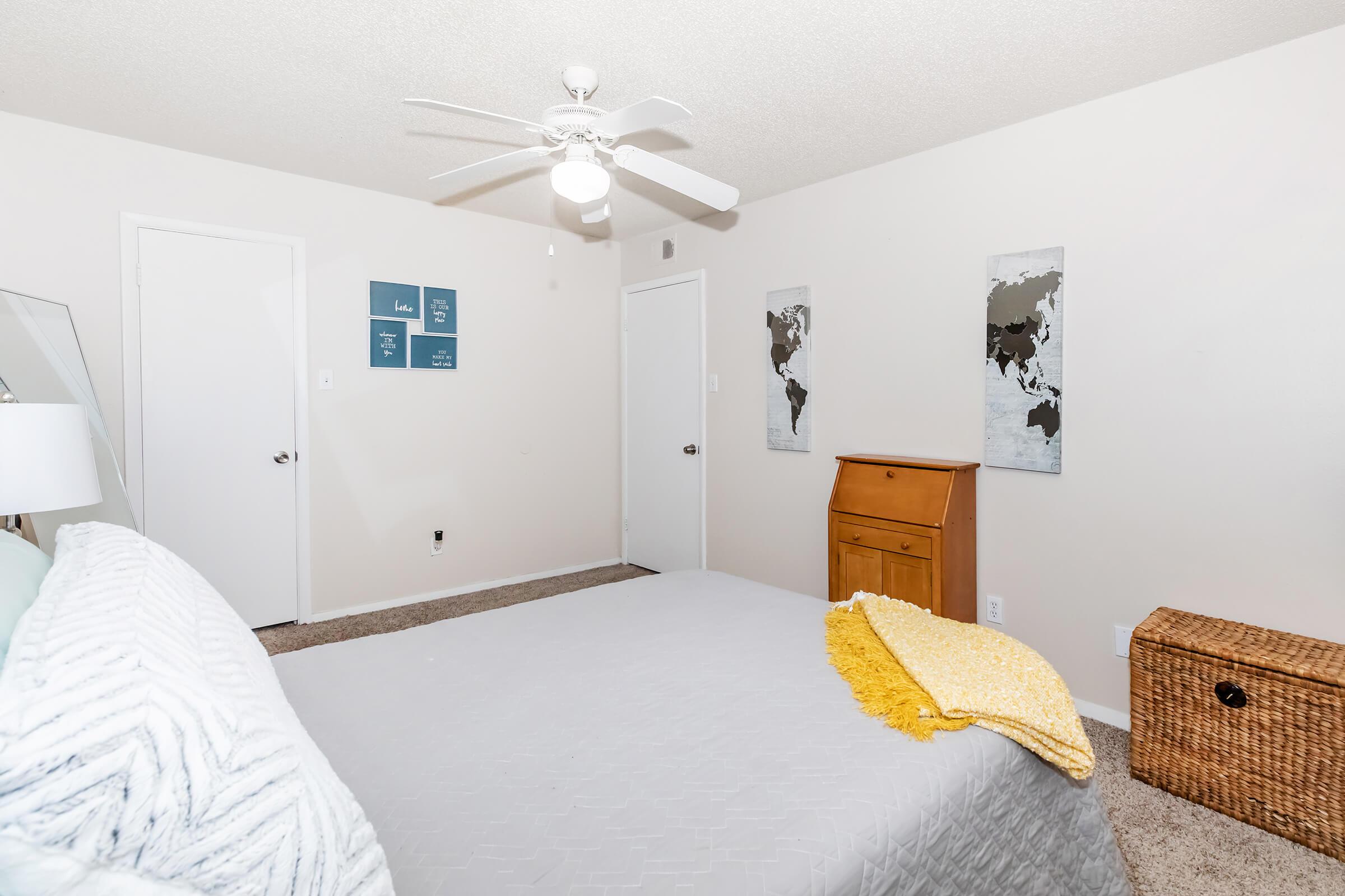 A neatly arranged bedroom featuring a light gray bedspread, a yellow throw blanket, a ceiling fan, and neutral-colored walls. There are two framed world map prints and decorative wall art consisting of quotes. The room includes a wooden dresser and a wicker basket for storage, with doors to a closet and another room visible.