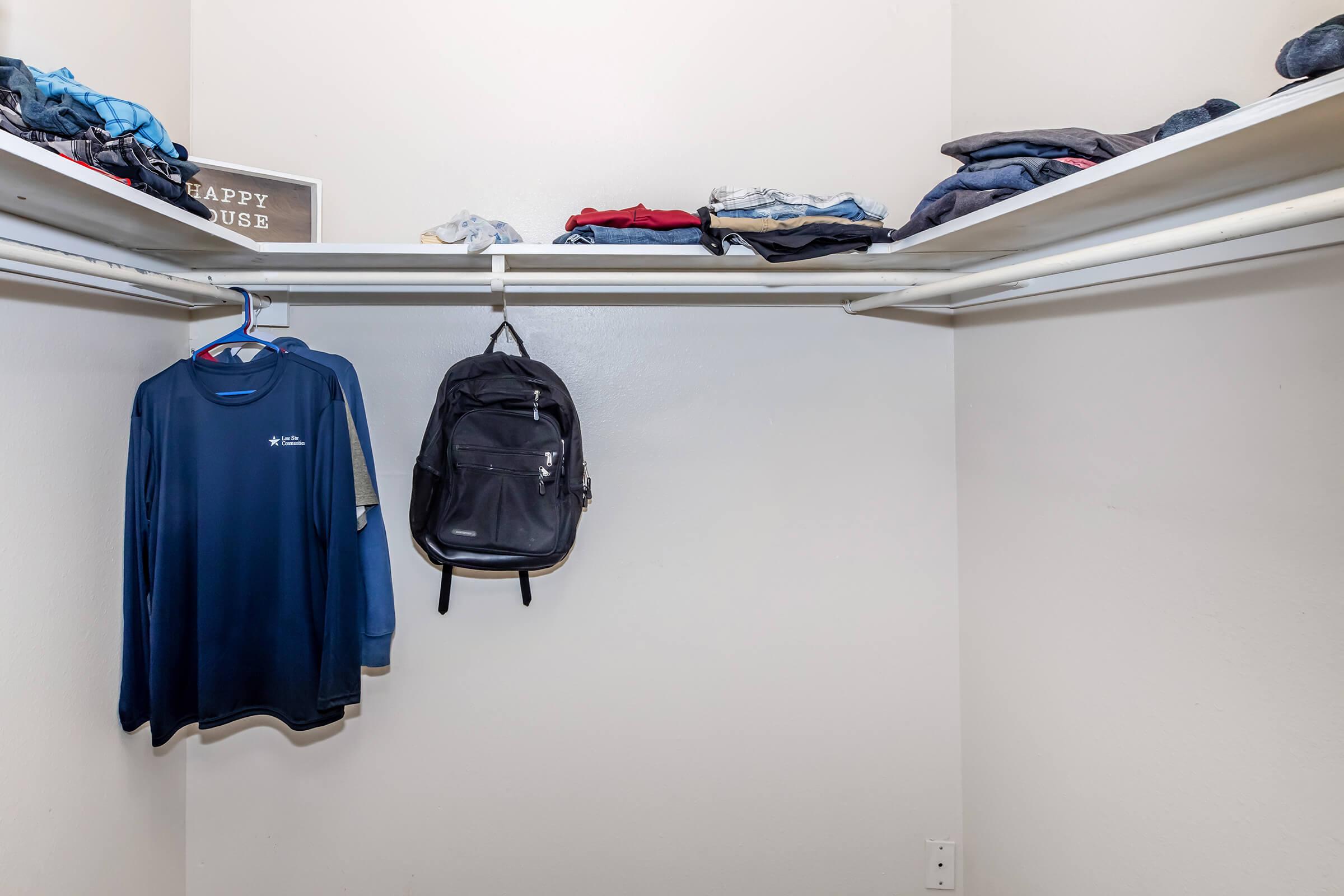 A neatly organized closet featuring a shelf with folded clothes, including denim and shirts, and a hanging black backpack on a clothes hanger. A small sign with the text "HAPPY HOUSE" is placed on the shelf. The walls are painted light gray, contributing to a clean and simple aesthetic.