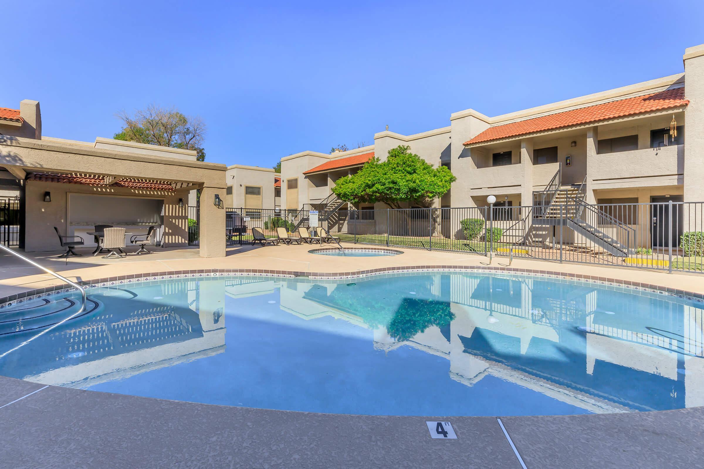 A clear blue swimming pool reflecting the sky, surrounded by a well-maintained courtyard featuring two-story apartment buildings with red-tiled roofs. There are lounge chairs and tables near the pool, along with neatly landscaped greenery, creating a relaxing outdoor space.
