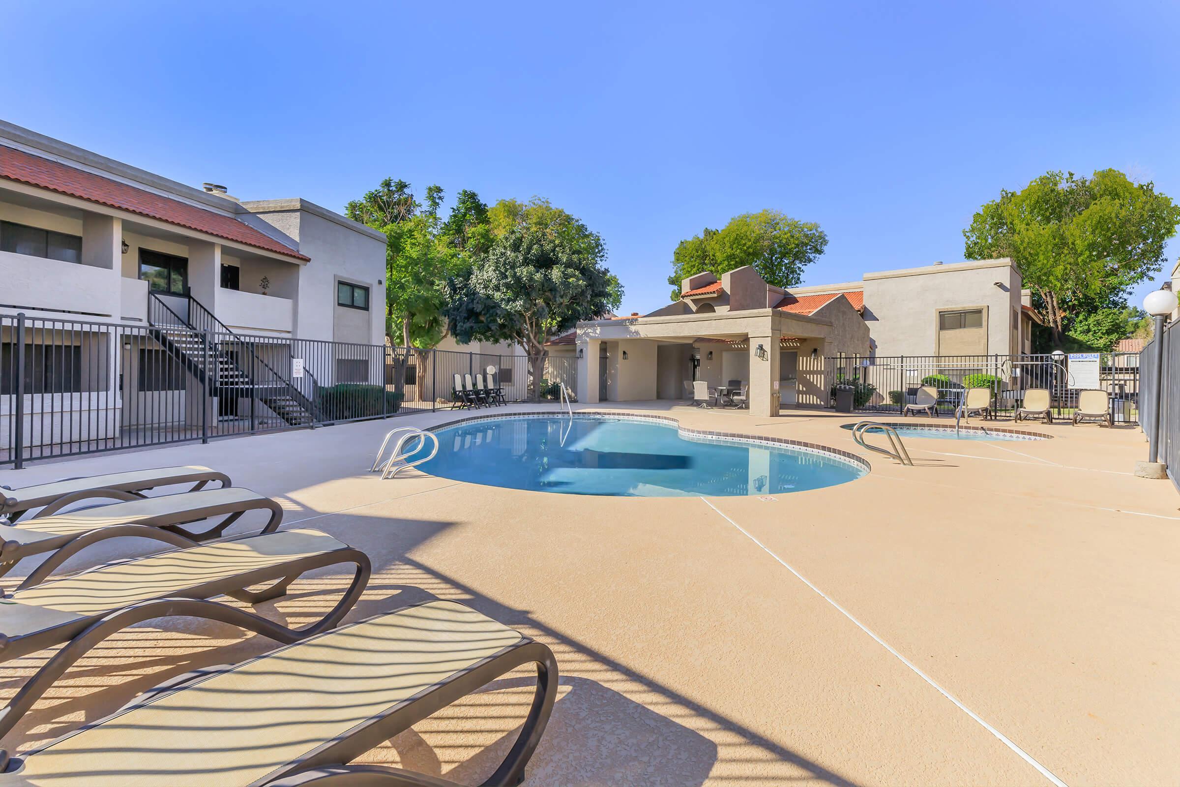 Swimming pool surrounded by lounge chairs and trees, with adjacent buildings in a sunny outdoor setting. The area features a covered patio with seating, creating a welcoming atmosphere for relaxation and leisure.