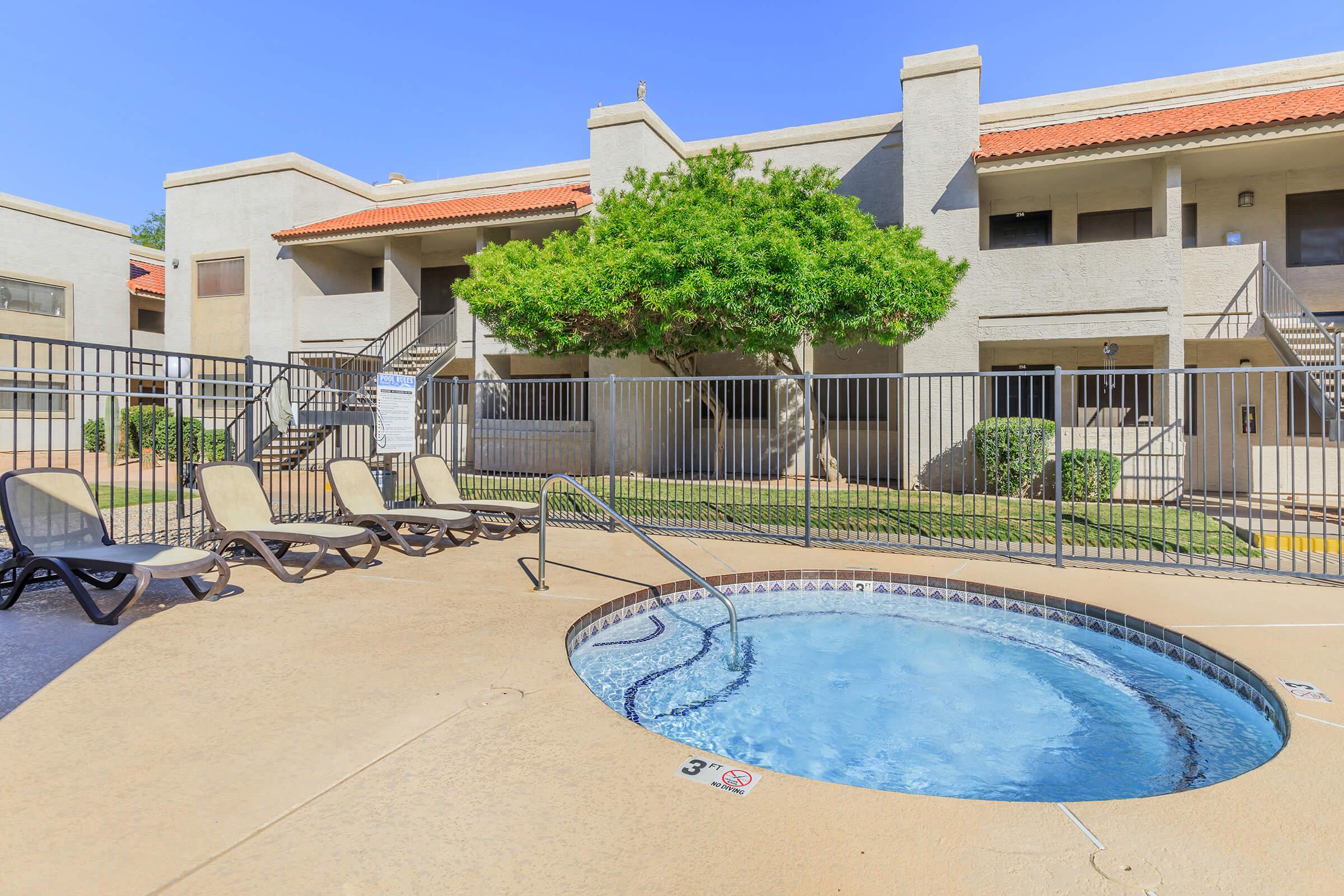 A small outdoor hot tub surrounded by a paved area with lounge chairs. In the background, a multi-story apartment building is visible, featuring a light-colored exterior and a red-tiled roof, with green shrubs and grass nearby. The scene is set under a clear blue sky.