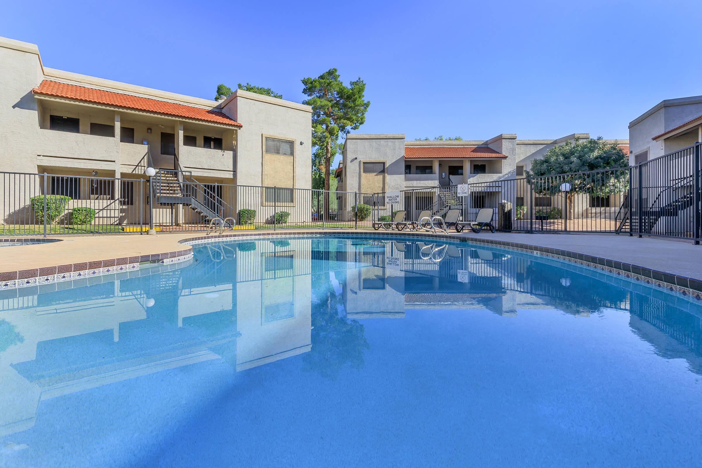 A clear swimming pool surrounded by lounge chairs, with modern apartment buildings and lush greenery in the background. The sky is blue, creating a relaxing atmosphere.
