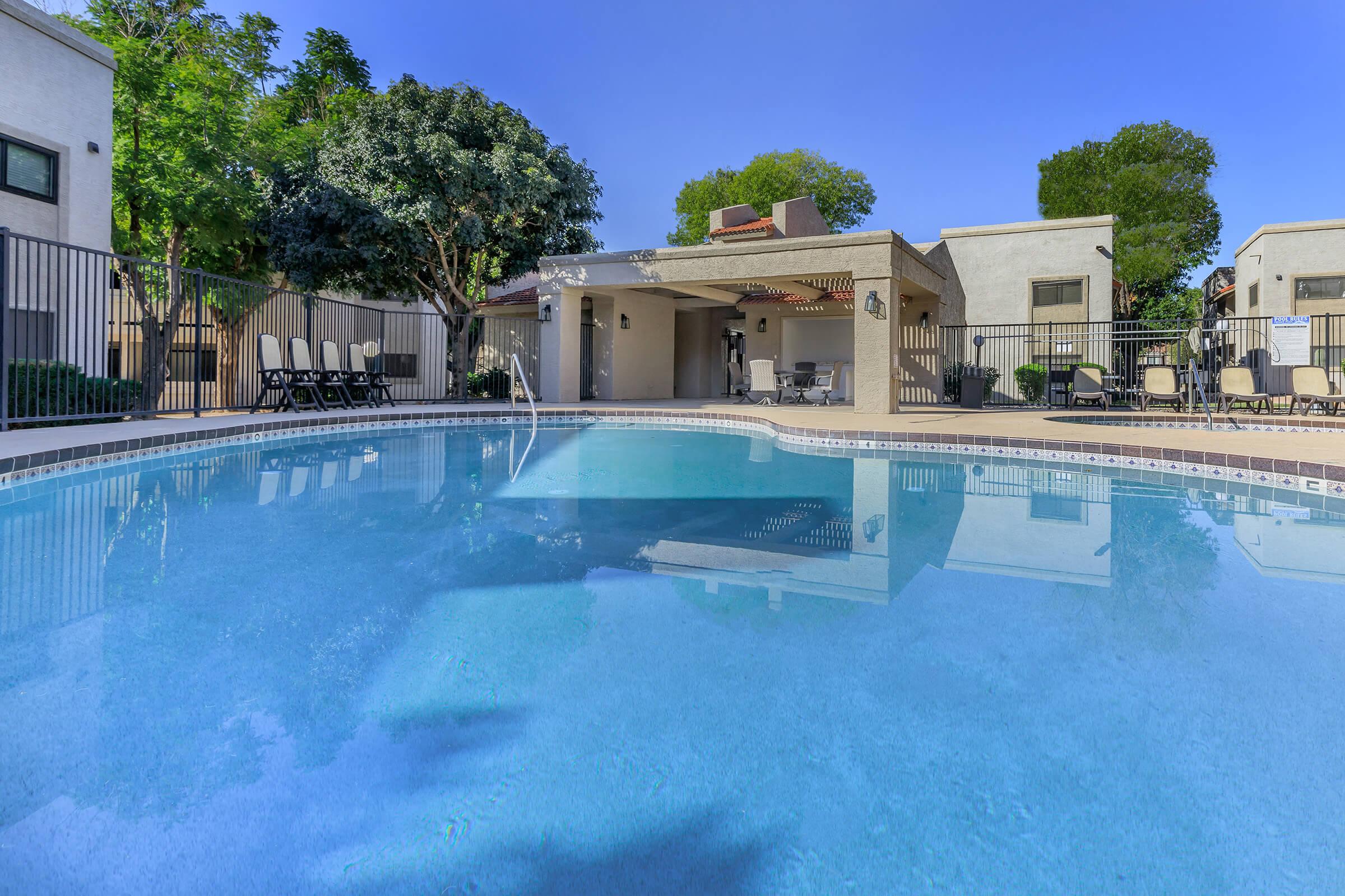 A clear blue swimming pool surrounded by lounge chairs and lush trees. A shaded area with a patio and seating is visible in the background, under a bright blue sky. The overall setting suggests a relaxing outdoor space, perfect for leisure and enjoyment.