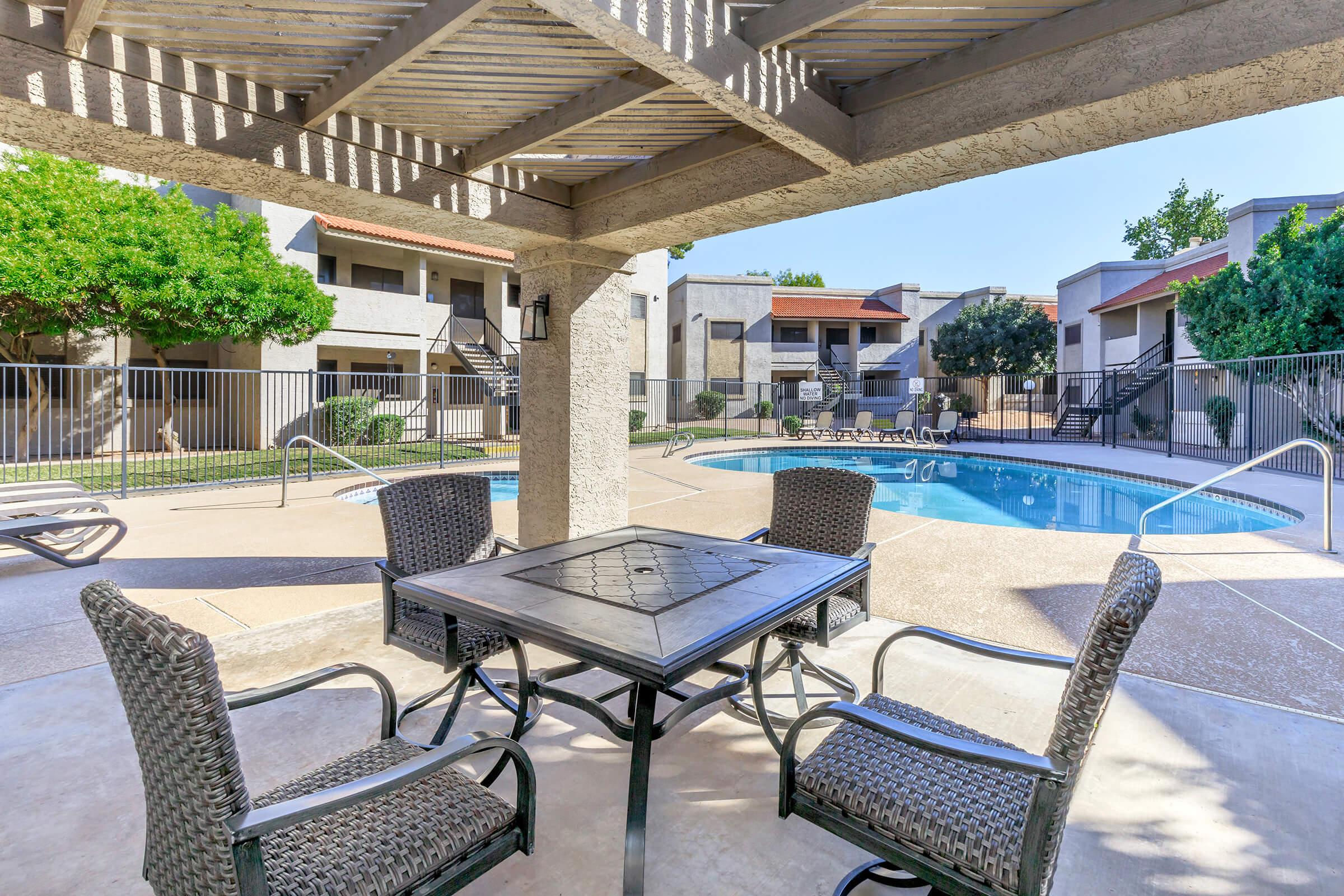 A view from a shaded patio area featuring a table and chairs overlooking a swimming pool surrounded by landscaped trees and apartment buildings. The scene is bright and inviting, ideal for relaxation or social gatherings.
