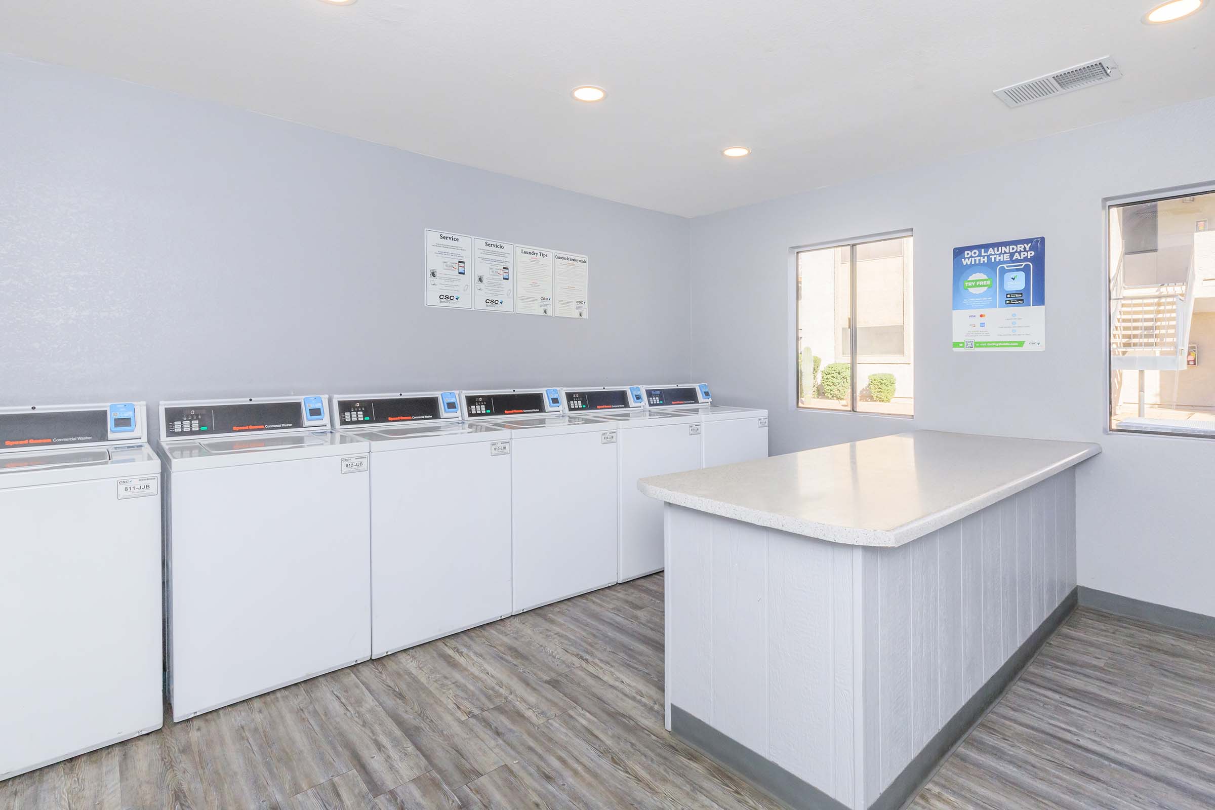 A clean and well-lit laundry room featuring multiple washing machines and a folding counter. The walls are painted in a light color, and there are windows allowing natural light to enter. Laundry-related posters and instructions are displayed on the walls.