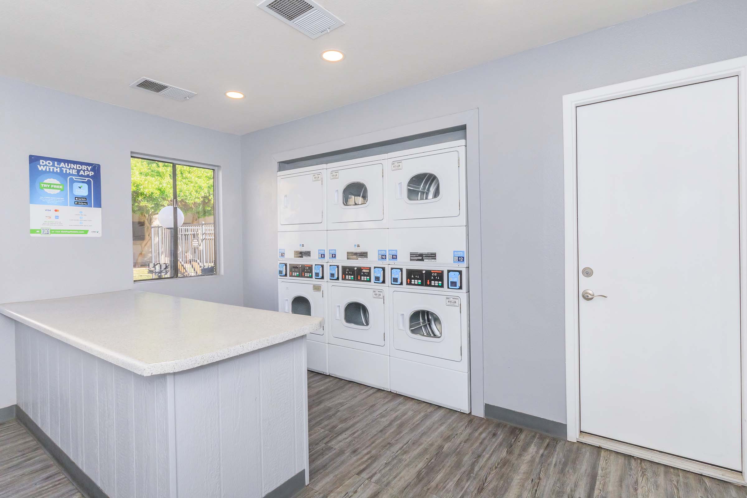 A clean, well-lit laundry room featuring several stacked washers and dryers against a gray wall. There's a countertop for folding laundry and a door leading to another area. A poster is visible on the wall with laundry service information. The flooring is a modern wood-look design.