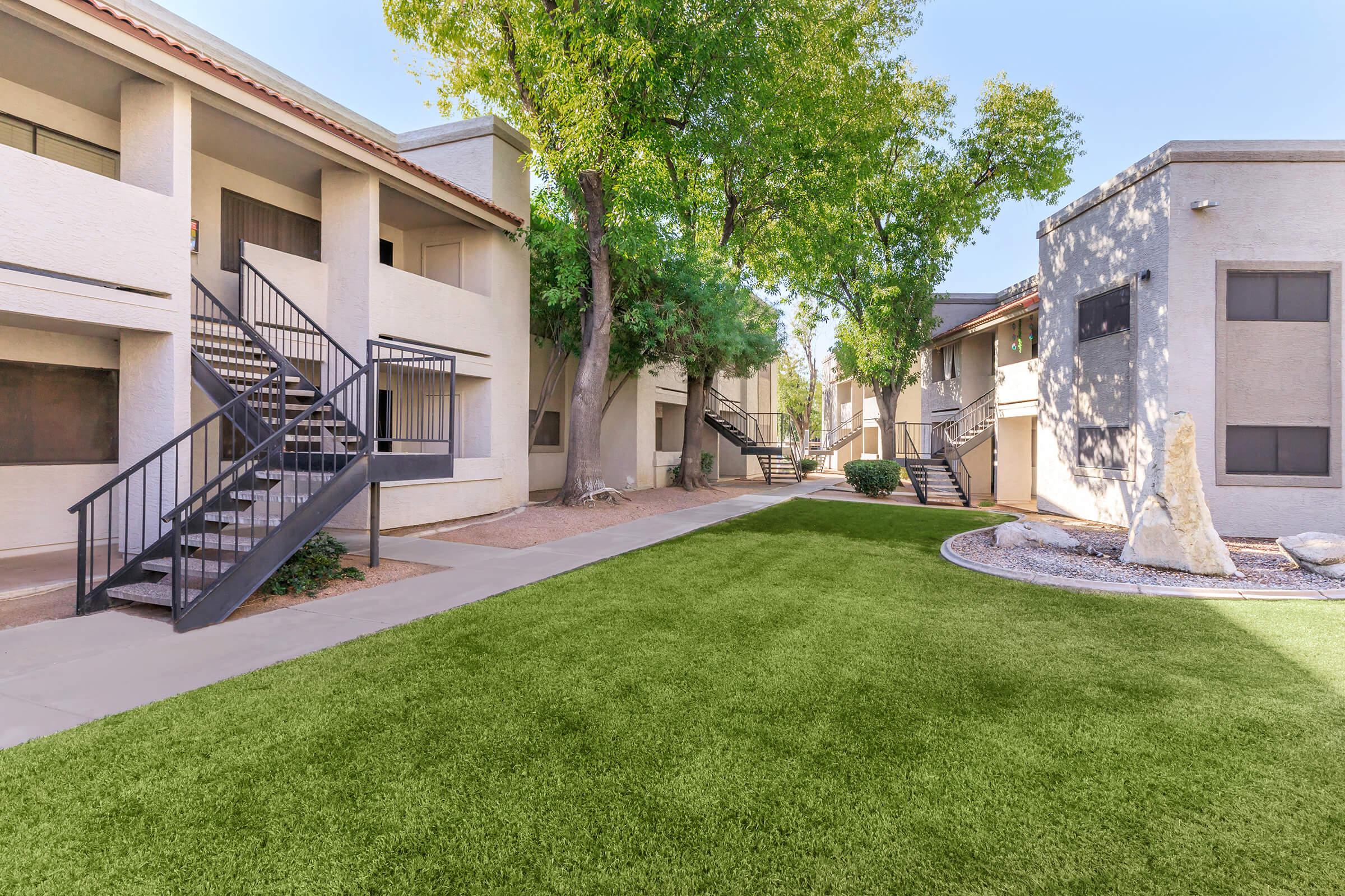 A landscaped courtyard of an apartment complex featuring green lawn, concrete pathways, and surrounding two-story buildings with exterior staircases. Trees provide shade, and decorative stones are placed in the landscape. Bright blue sky adds to the inviting atmosphere.