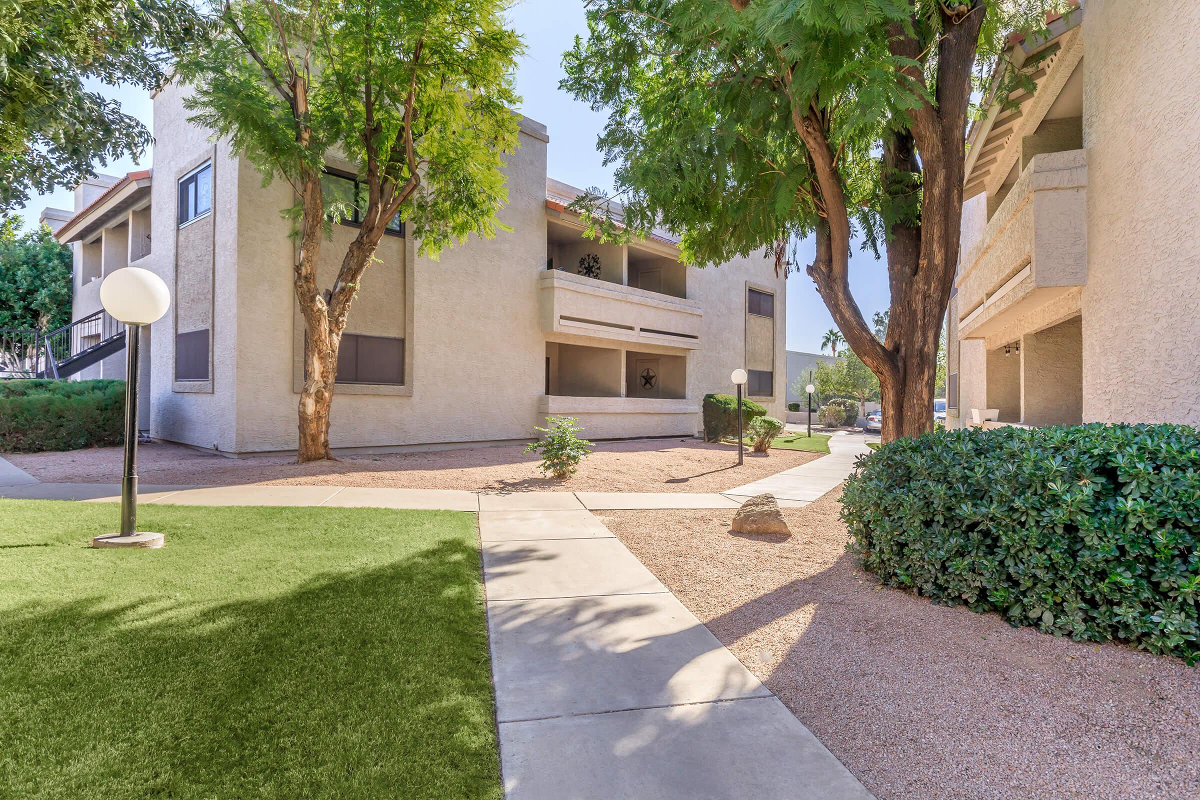 A well-maintained outdoor area featuring two-story apartment buildings surrounded by lush green trees and shrubs. A paved walkway meanders through the space, while decorative light fixtures add to the ambiance. The grounds are neatly landscaped, with vibrant vegetation contrasting against the buildings.