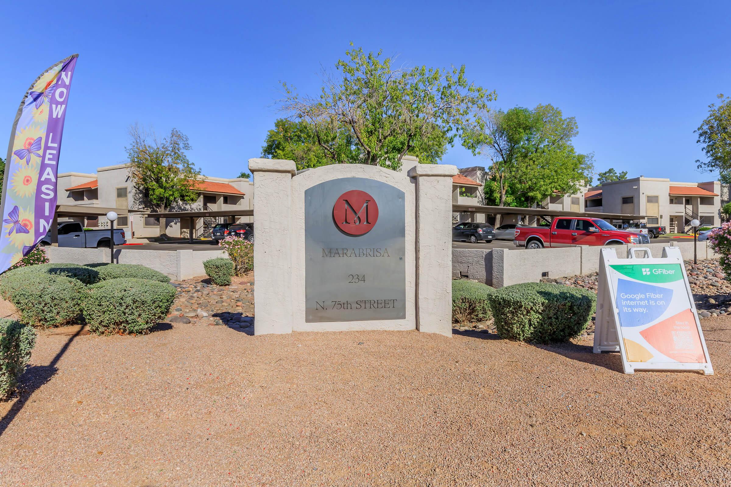 Sign for "Mariposa A" located at 2934 N. 76th Street, featuring a prominent logo. Surrounding the sign are well-maintained shrubs and desert landscaping. In the background, a multi-unit building can be seen. A colorful banner reads "Now Leasing," and there is a sign indicating "Google Fiber is coming."