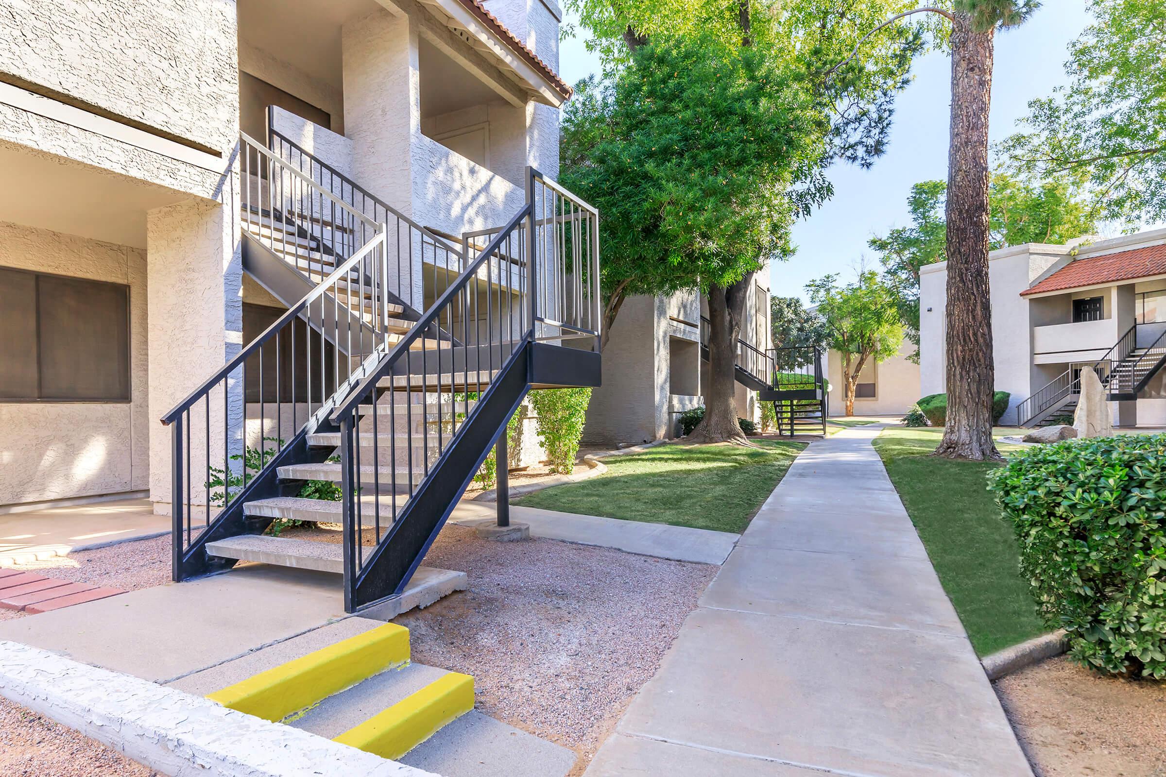 A clear path leads through a residential complex, featuring buildings with staircases. Lush greenery and well-maintained lawns are visible, along with a paved walkway. The scene is sunny, highlighting the architectural design and landscaping of the area.
