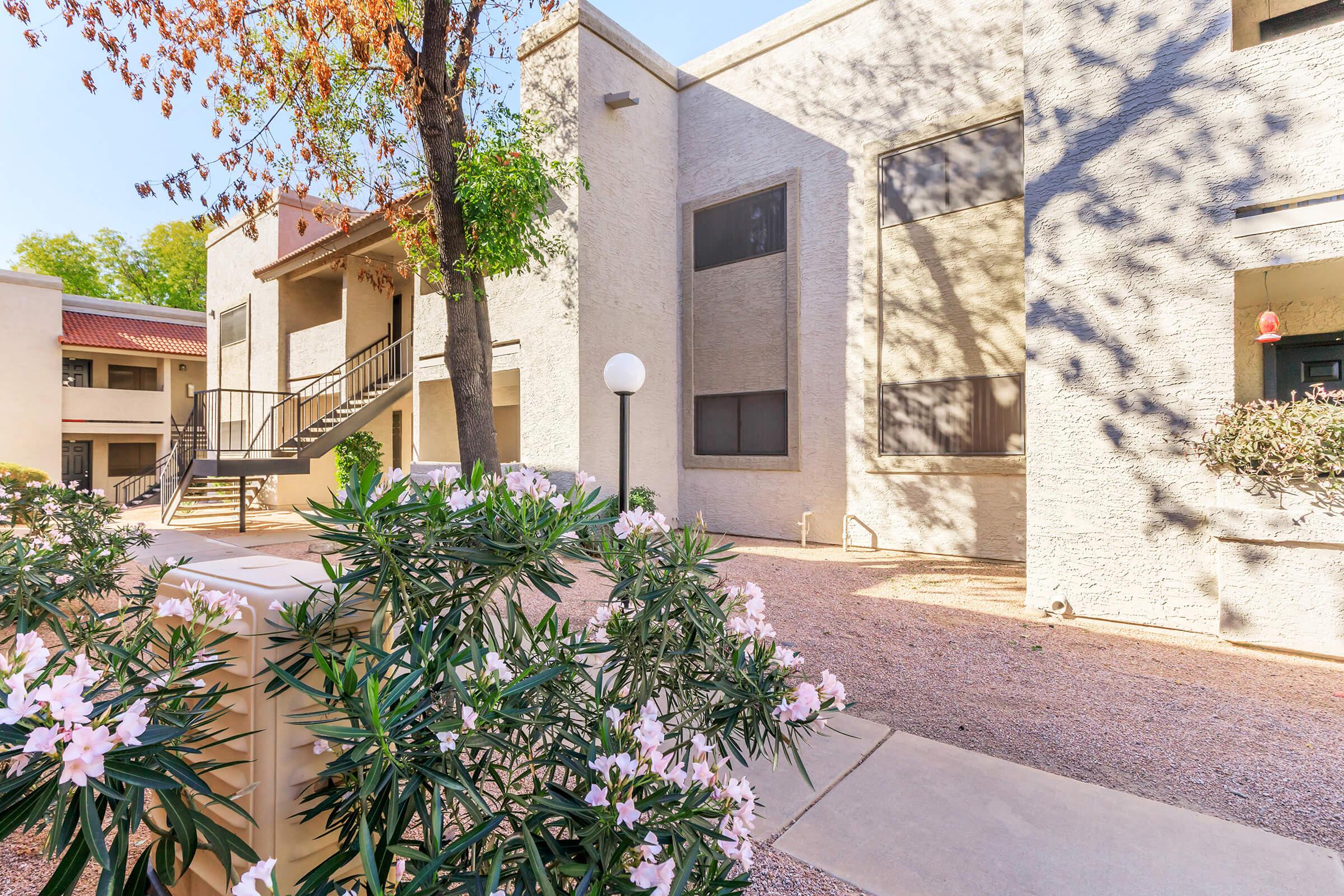 A well-lit outdoor area featuring a low-rise apartment building with textured gray walls. In the foreground, there are flowering bushes and a pathway leading up to the building. Tall trees cast dappled shadows, and a set of stairs leads to a second-floor entrance. The setting is tidy and visually appealing.