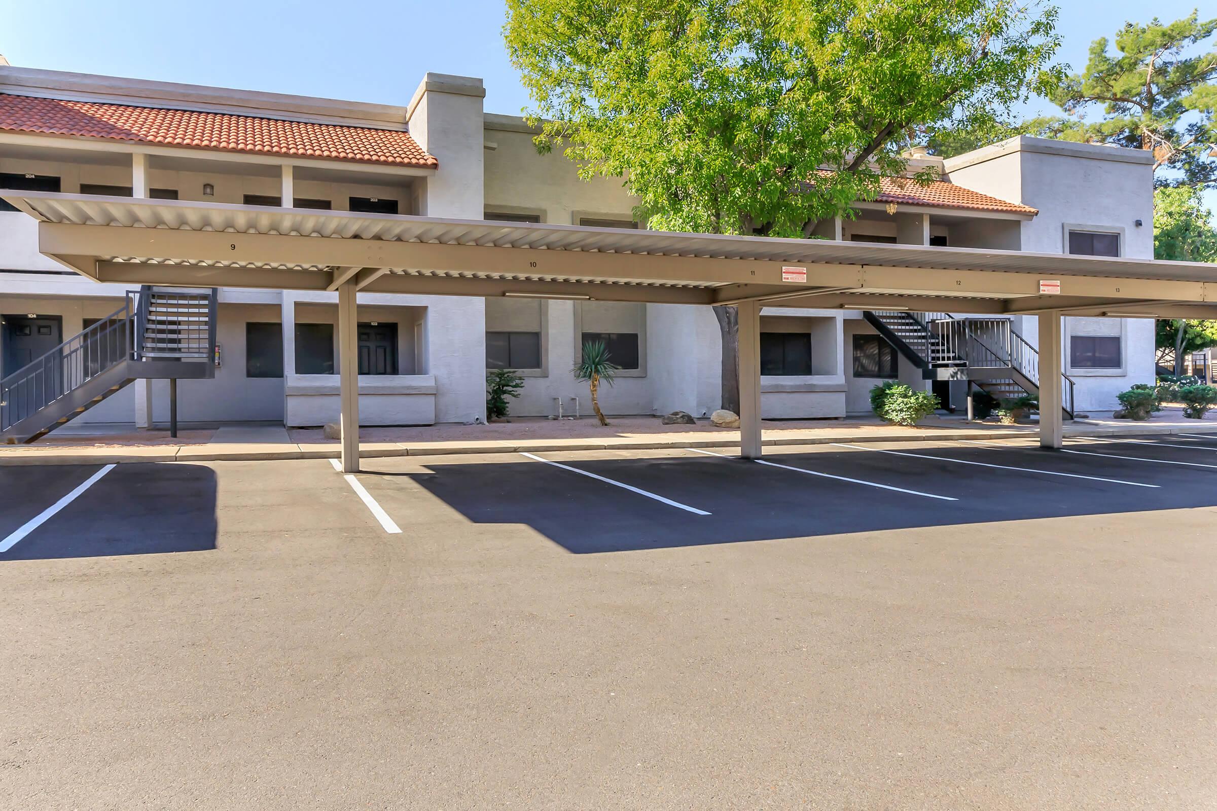 Covered parking spaces in front of a two-story apartment building, featuring a light-colored exterior with a red-tiled roof. A staircase leads to the second floor, and a large tree provides shade nearby. The parking area is empty with clearly marked spaces.