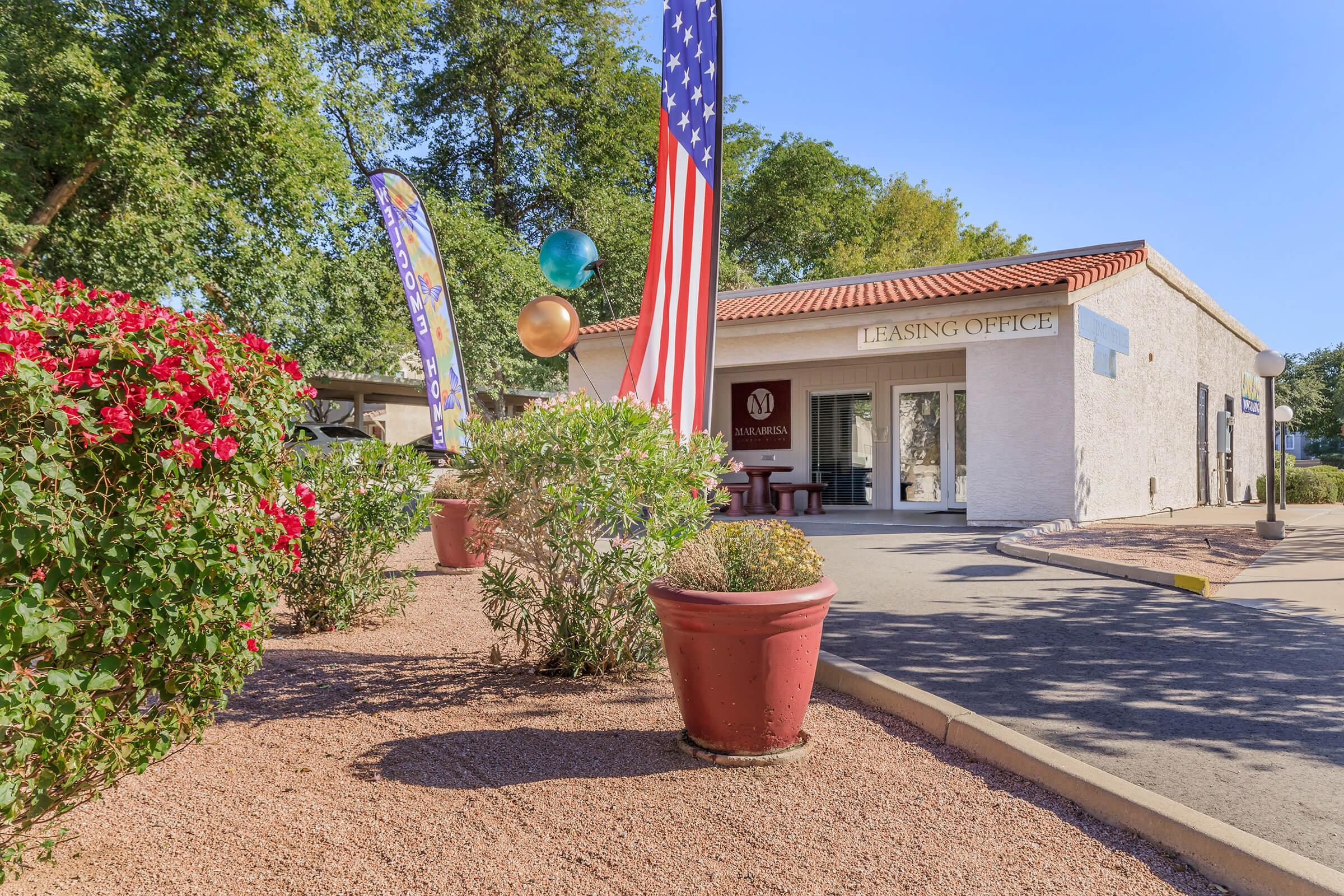 A leasing office building with a welcoming exterior, featuring a sign that reads "Leasing Office." The entrance is decorated with colorful balloons and American flags. Surrounding the office are potted plants and blooming flowers, set against a clear blue sky and green trees in the background.
