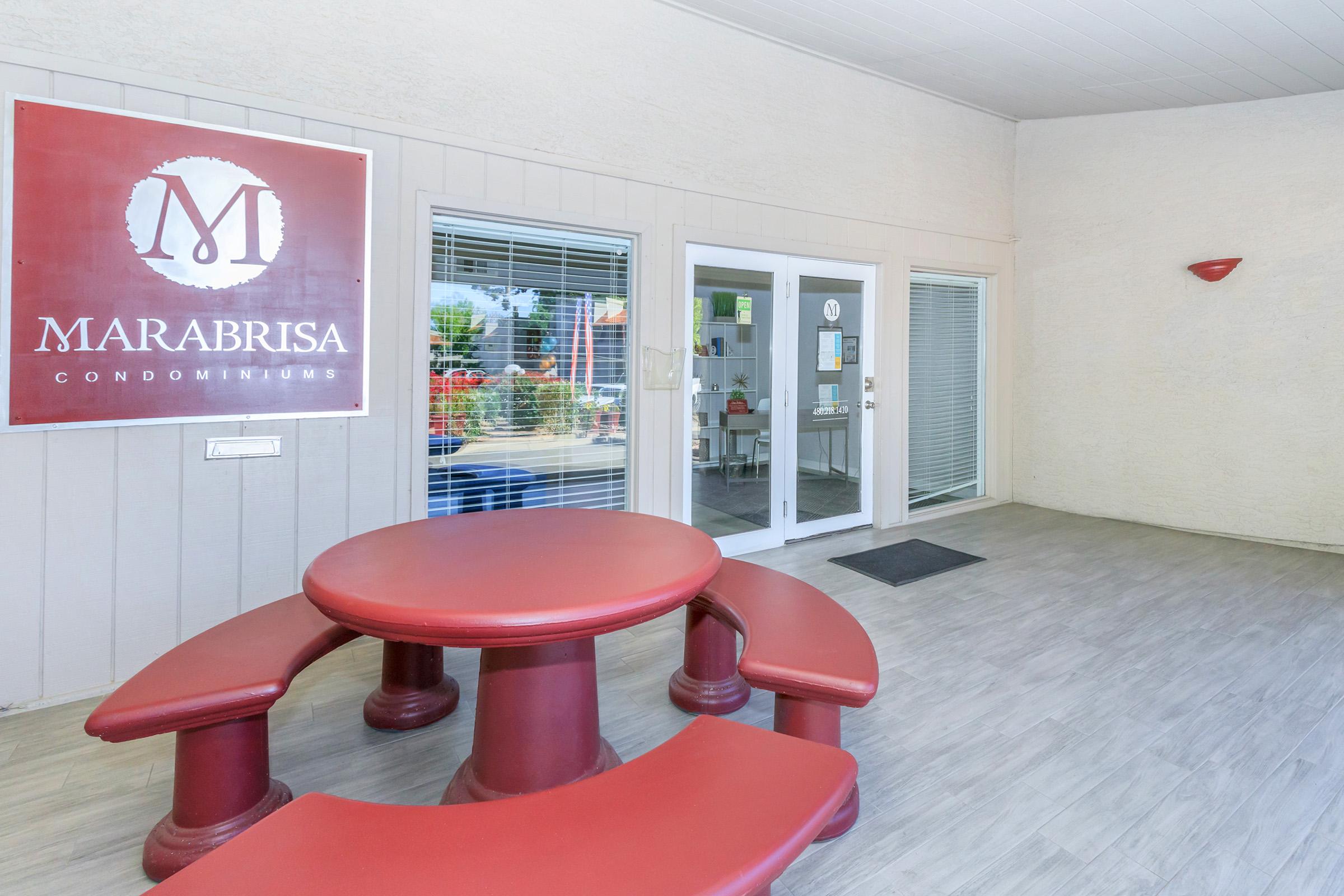 Exterior view of the Marabrisa Condominiums entrance, featuring a red sign with the name prominently displayed. In front, there is a round dining table with benches, set against a light-colored wall and large windows. The area appears clean and welcoming, with greenery visible outside.