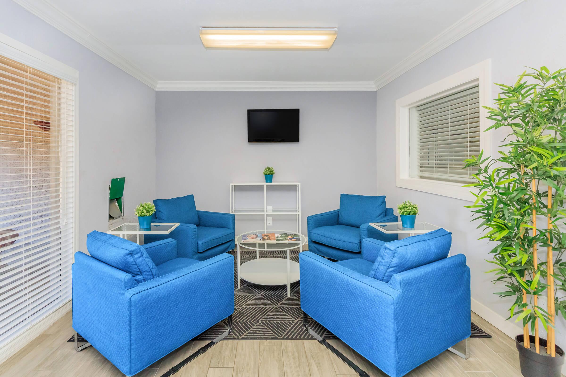 A cozy lounge area featuring four blue upholstered chairs arranged around a glass coffee table. A small plant decorates the table, and there is a flat-screen TV mounted on the wall above. The space is brightened by natural light from window blinds and has a subtle, modern aesthetic with a bamboo plant in the corner.