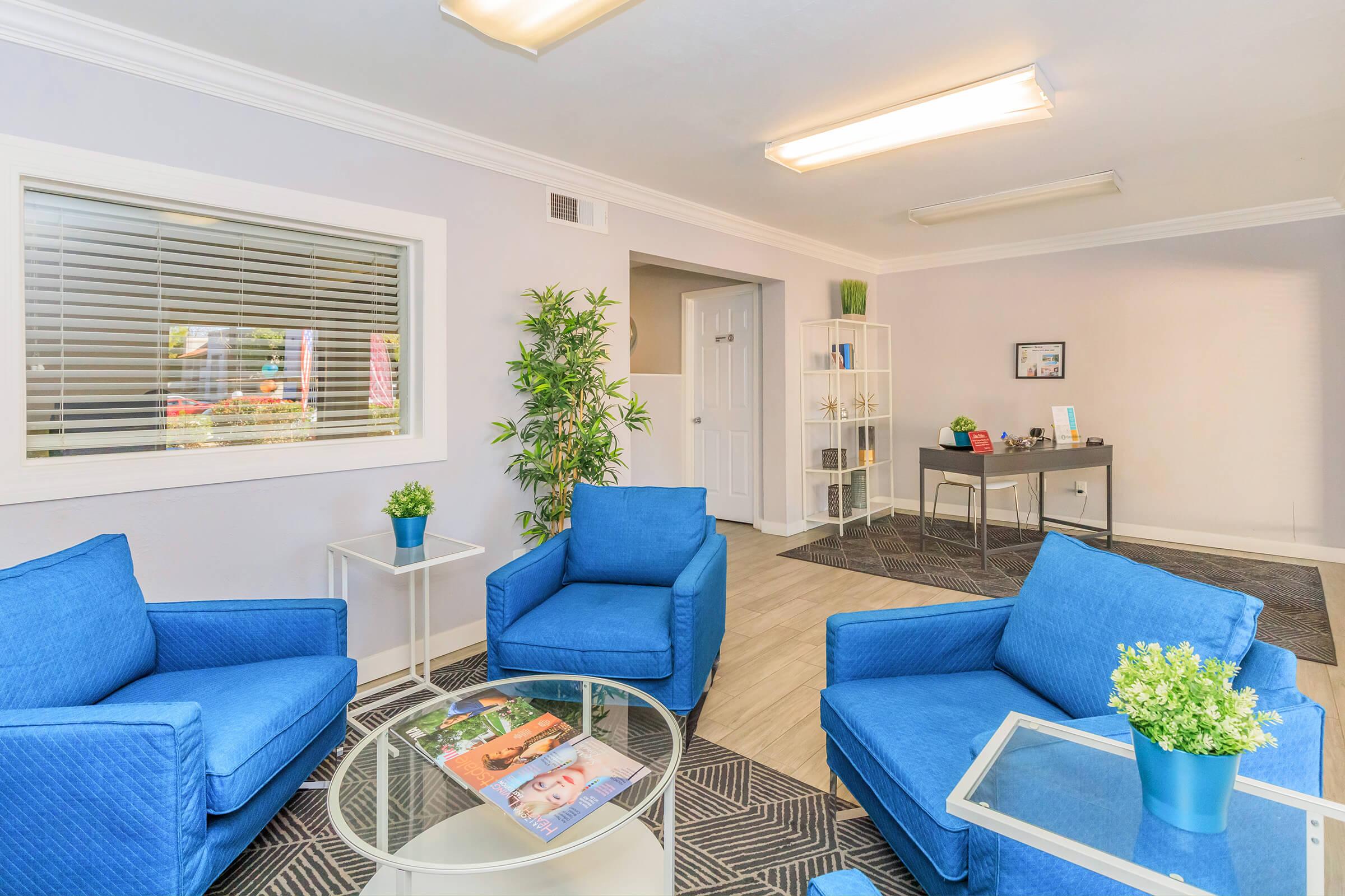 A modern waiting area featuring two blue armchairs, a glass coffee table with a magazine, and a small plant. Light-colored walls and flooring create a bright atmosphere, while a black console table holds decorative items. Windows allow natural light, enhancing the inviting feel of the space.