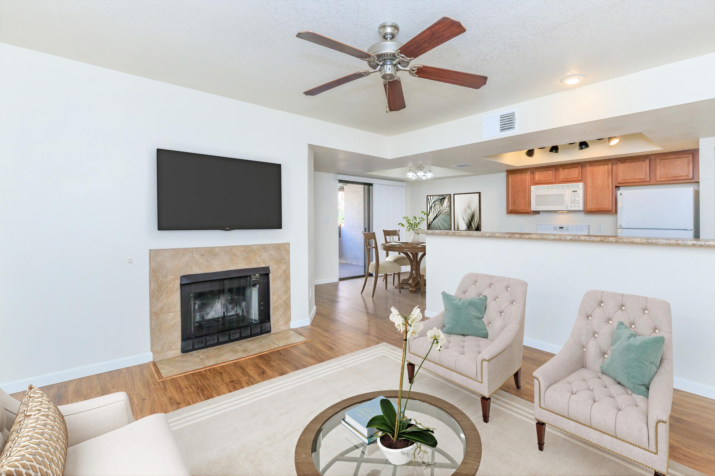 A cozy living room with a light-colored sofa and two tufted armchairs. A round coffee table with a potted plant sits in the center. A flat-screen TV is mounted on the wall above a stone fireplace. In the background, a dining area with a table and chairs is visible, along with a kitchen space. Natural light streams in from nearby windows.