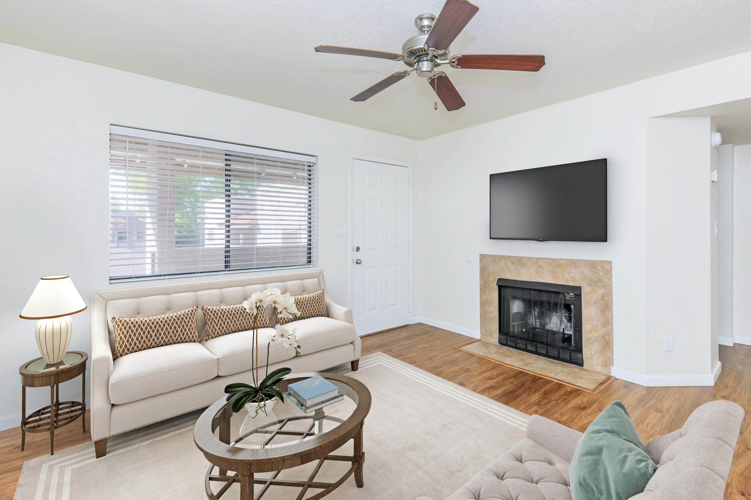 A bright and airy living room featuring a light-colored sofa with decorative pillows, a coffee table with a book, and a cozy fireplace. A ceiling fan hangs above, and large windows provide natural light. A flat-screen TV is mounted on the wall opposite the seating area. The decor is modern and inviting.