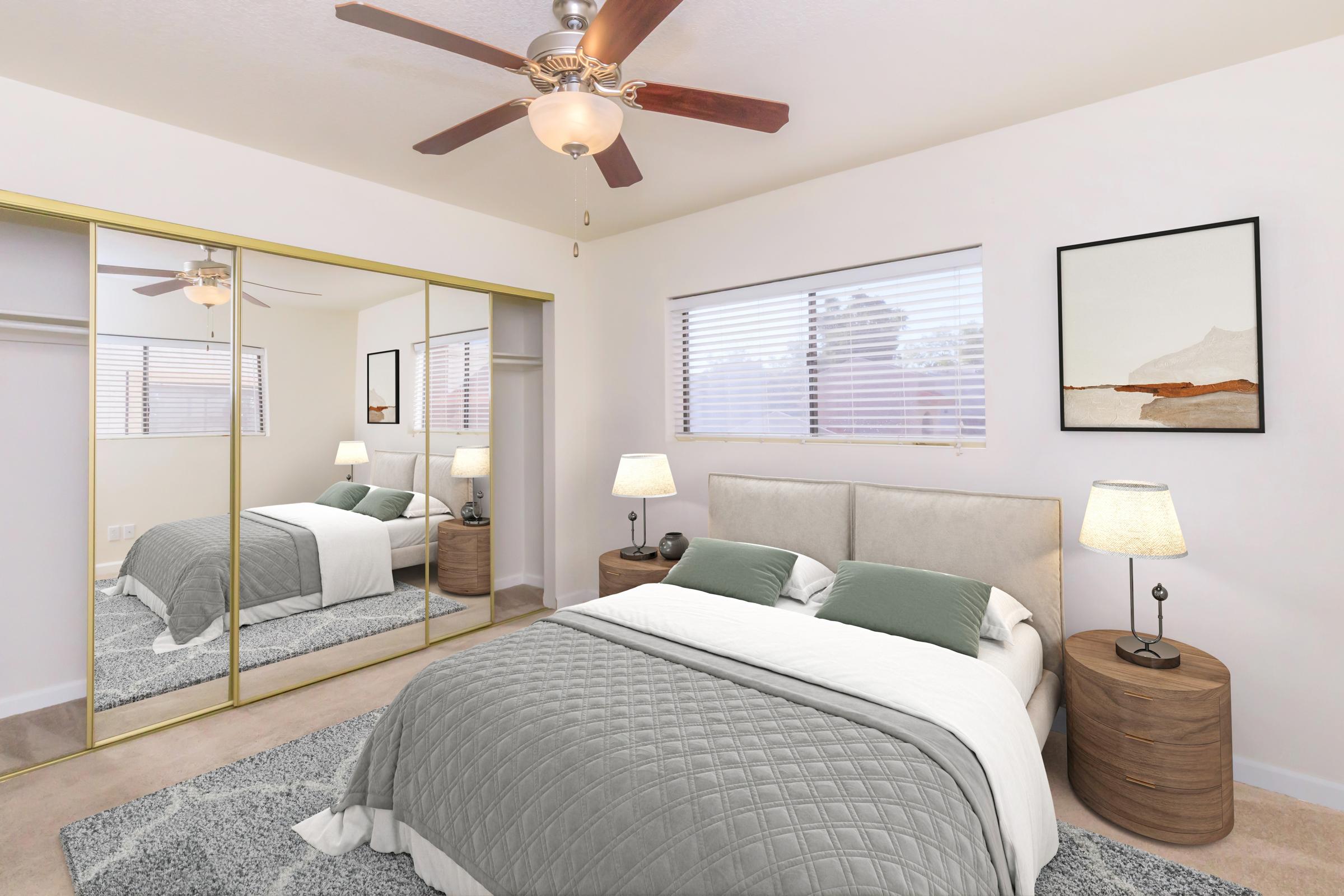 A modern bedroom featuring a queen-sized bed with gray bedding, two bedside lamps on wooden tables, a large mirror closet with sliding doors, and art on the wall. The room has a ceiling fan and natural light filtering through the windows, creating a cozy, inviting atmosphere.