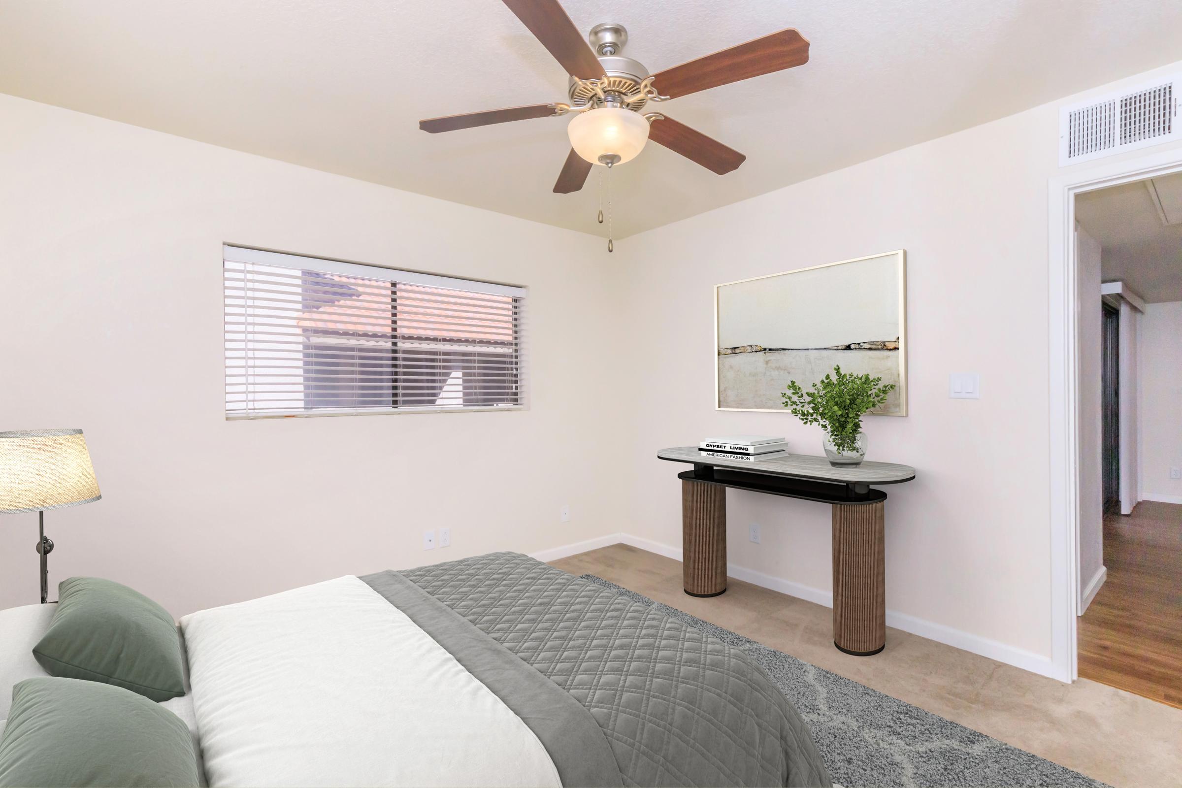 A modern bedroom featuring a light-colored wall, a large window with blinds, a ceiling fan, a neatly made bed with green and gray bedding, a bedside lamp, and a small console table adorned with a plant and a decorative piece. The room has a clean and minimalist design.