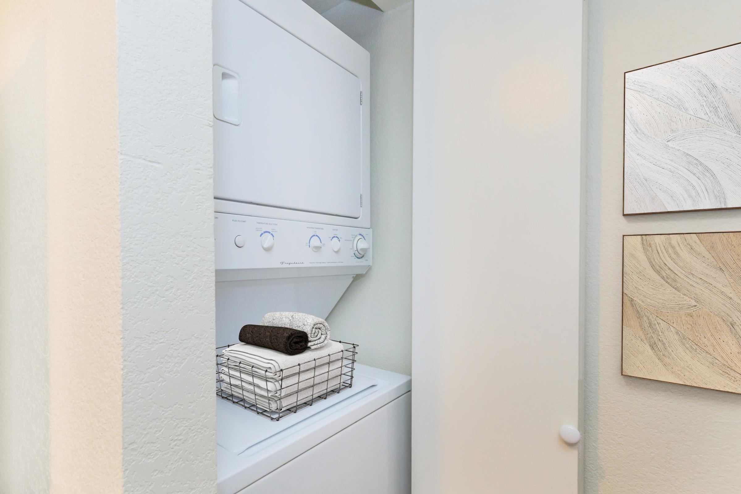 A compact laundry nook featuring a stacked washer and dryer. A wire basket containing neatly folded towels sits on the dryer, and the space is adorned with light-colored walls and decorative panels nearby, creating a clean and organized laundry area.