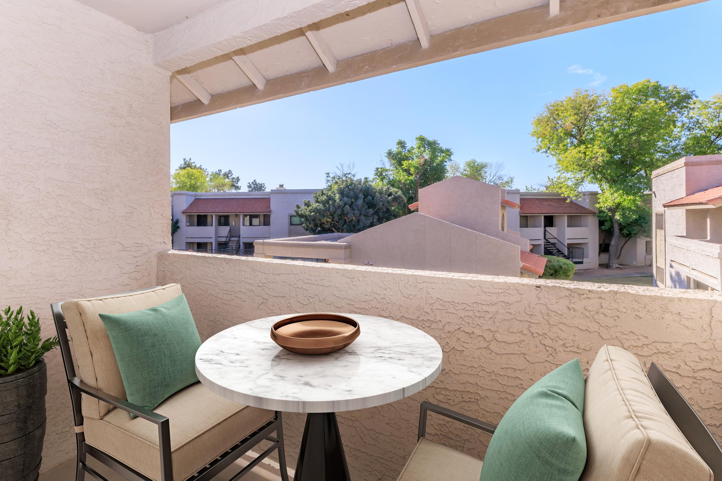 A cozy balcony featuring a round marble table with a brown bowl on top, surrounded by two modern chairs with green cushions. In the background, a view of neighboring buildings and trees under a clear blue sky.