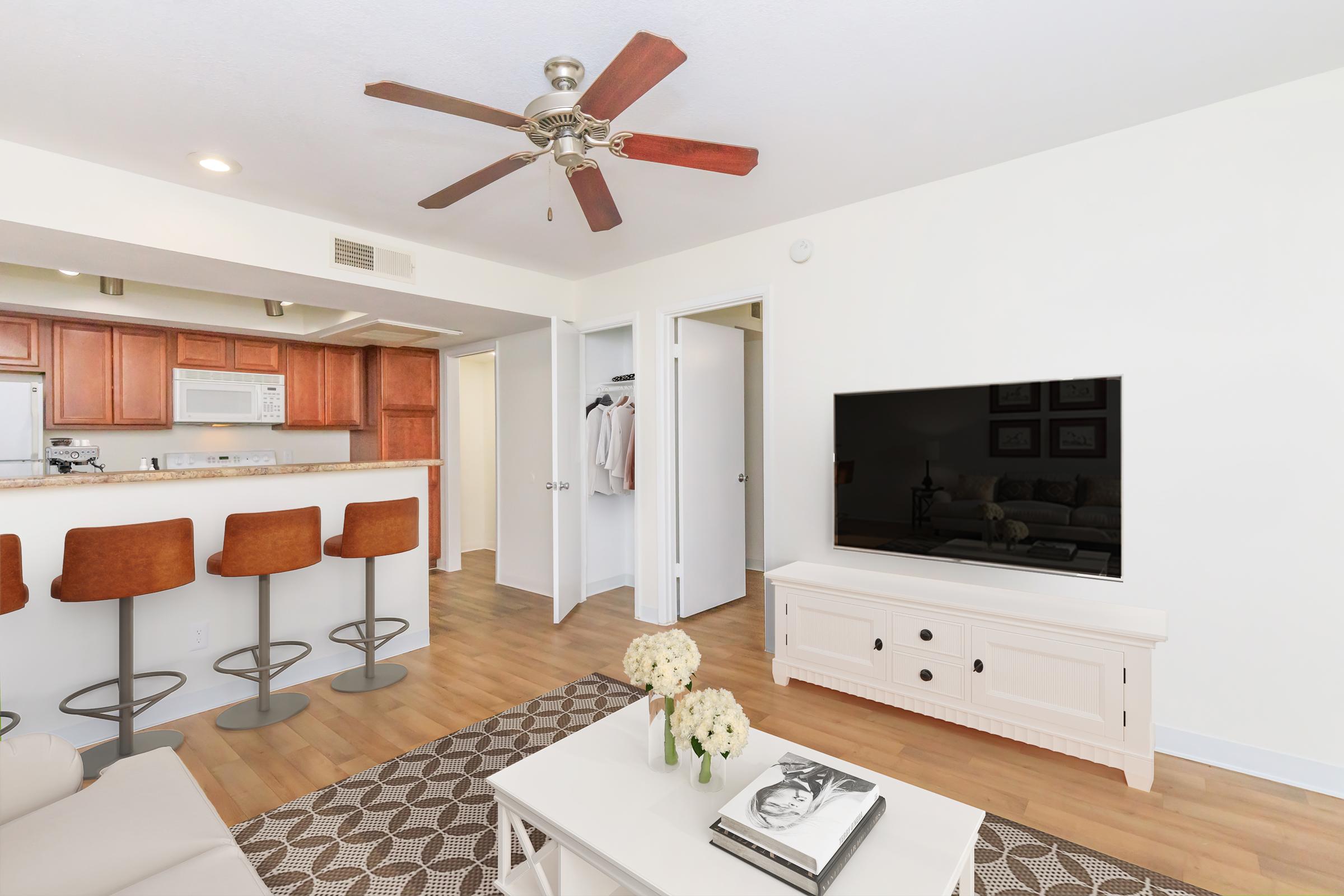 Interior view of a bright living room featuring a white coffee table with flowers, a flat-screen TV on a light wood console, and a beige couch. In the background, there's a bar area with three orange stools and a kitchen space with wooden cabinets. The room has a ceiling fan and neutral-colored walls.